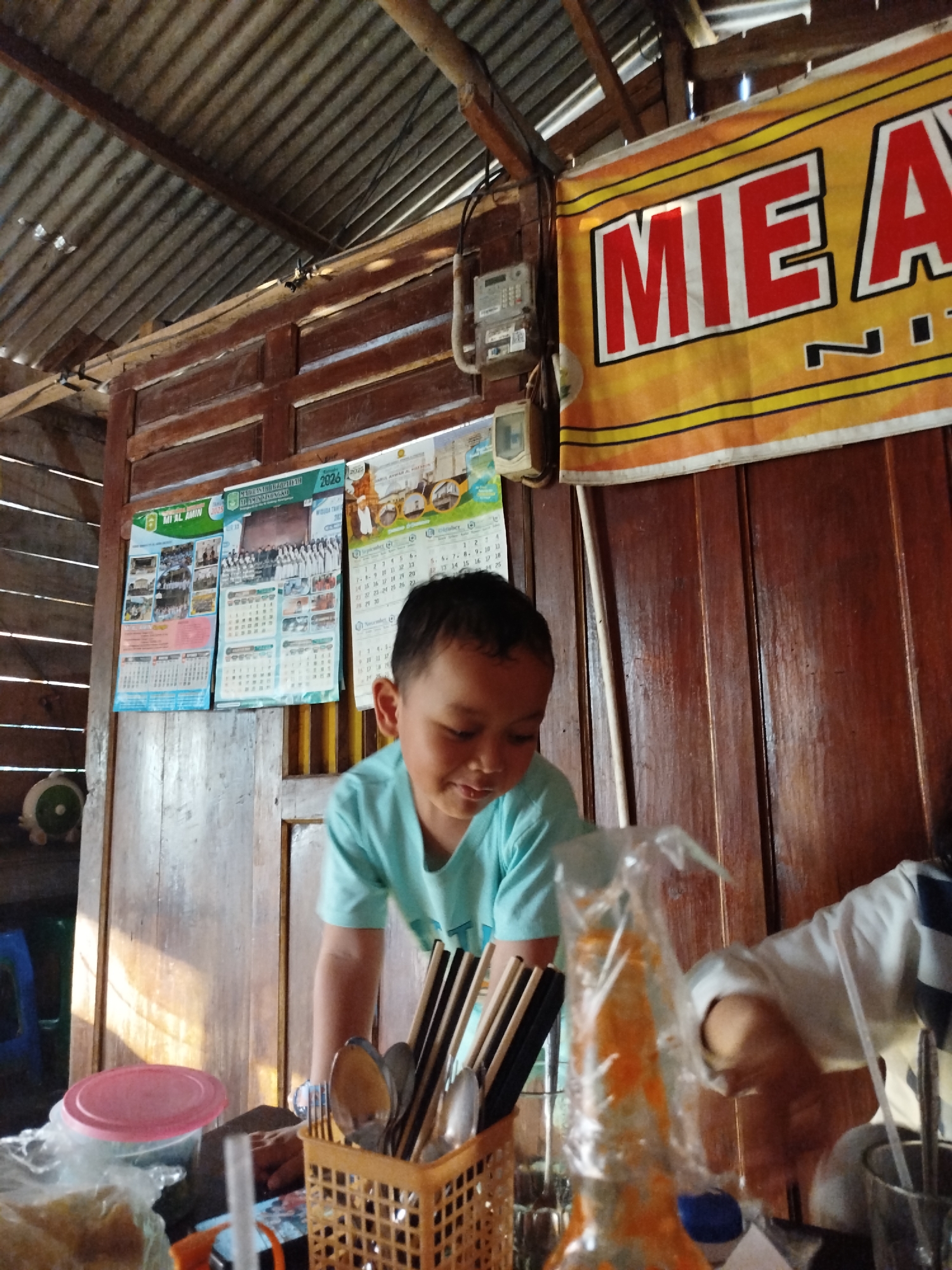 The image depicts an indoor scene, likely a food stall in Pojok, Indonesia, given the signage and general ambiance. A young boy, wearing a light blue t-shirt, is leaning over a counter. He has dark hair and is smiling. In front of him, on the counter, are various items including a plastic basket filled with chopsticks and utensils, a pink lid from a container, and a plastic-wrapped bottle containing an orange liquid. Behind the boy, a yellow banner with red lettering reads "MIE A," suggesting it is advertising noodles. Electrical meters are visible on the wooden wall. Several calendars, dated 2023 and 2025, are also hanging on the wall. The overall environment appears rustic, with a corrugated metal roof supported by wooden beams and wooden walls. Another person, wearing a white shirt with dark stripes, is partially visible to the right, interacting with the scene.
