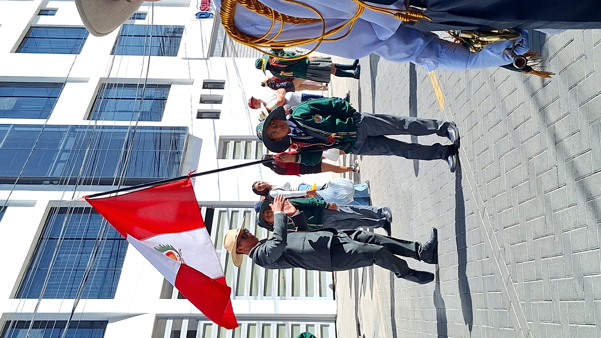 Cerro Colorado, Peru. A parade on a paved street next to a building with a glass facade. A man in a dark suit and straw hat stands next to a flag of Peru. Behind him are students in school uniforms with dark blazers and hats. Some are carrying flagpoles. A line of people in various outfits, including one with a kilt, is visible further back.