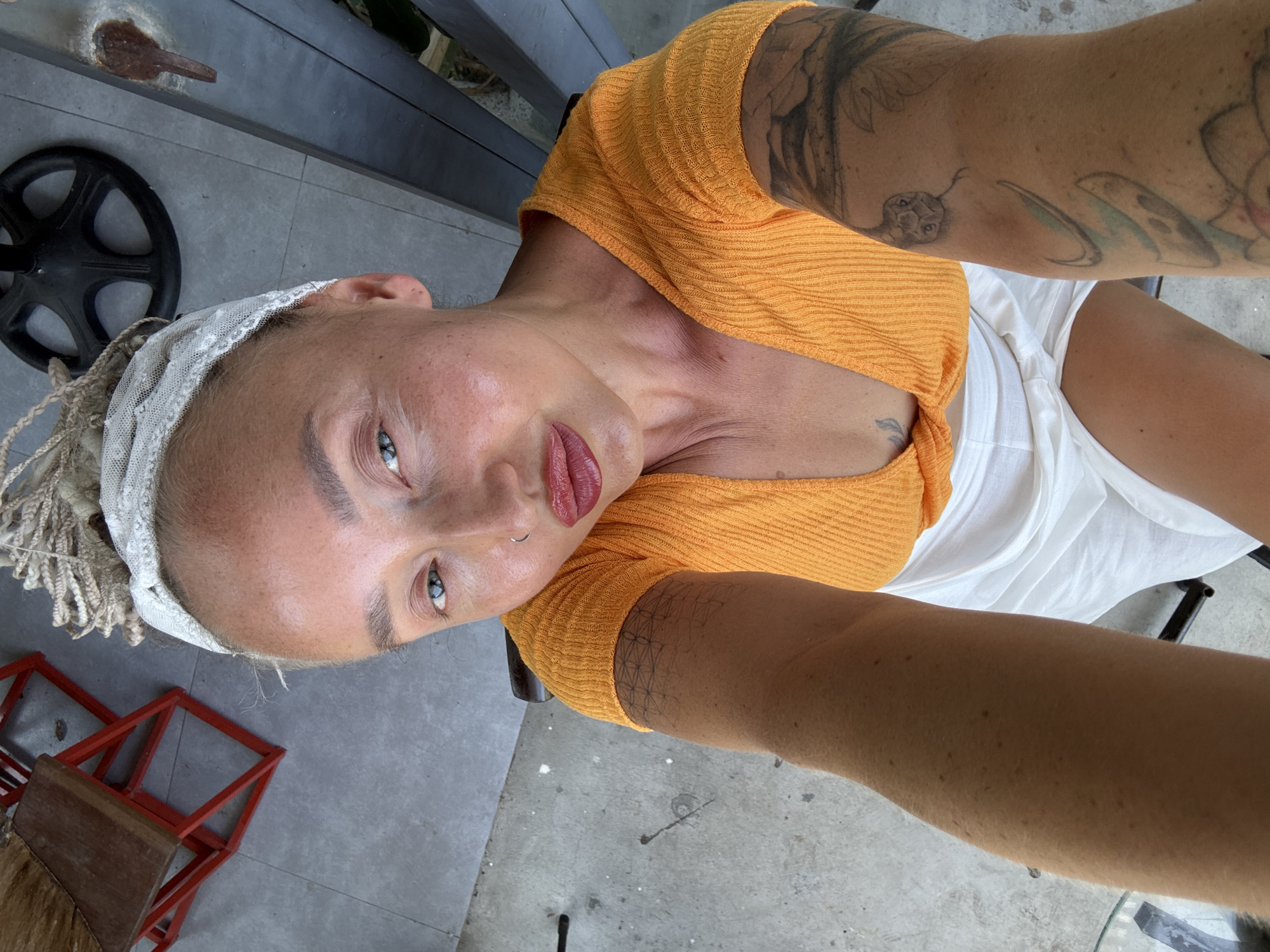 A woman with blonde dreadlocks, a white lace headband, and a silver nose ring is taking a selfie. She is wearing an orange ribbed short-sleeved top and a white skirt. Her arms are visible, showing tattoos. The background appears to be an outdoor setting, possibly a cafe or restaurant patio, with a grey concrete floor, a red metal chair with a wooden seat, and some dark vertical beams. The image is taken from a high angle, looking down at the woman. The location is identified as Bac Nha Trang Ward, Vietnam.
