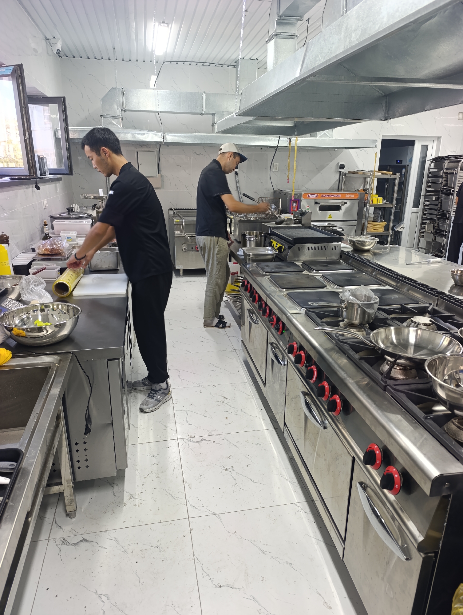 In an indoor commercial kitchen with white marble-patterned tile flooring and a metallic ceiling, two men in dark chef's uniforms are preparing food. The man on the left, facing the camera and wearing gray sneakers, is holding a roll of yellow plastic wrap. The man on the right, wearing gray pants and white sneakers, is facing away from the camera and appears to be cooking at a stainless steel range with multiple burners and knobs. A large exhaust hood extends across the kitchen ceiling above the cooking area. Various kitchen equipment, including a large stainless steel bowl, pots, pans, and a griddle, are visible on the counters and the range. To the left, a window with an open sash reveals a glimpse of an outdoor scene with trees. A security camera is mounted on the wall above the window. The overall environment suggests a professional culinary setting.