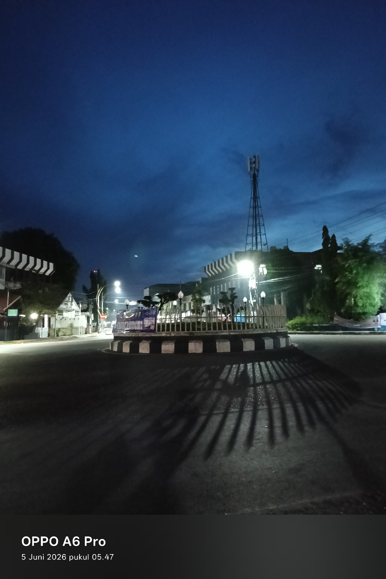 The image captures a quiet urban street scene at dawn in Lhokseumawe, Indonesia. The sky is a deep, dark blue with hints of lighter clouds, indicating the early morning hours, confirmed by the timestamp "5 Juni 2026 pukul 05:47" (June 5, 2026, 05:47 AM). No people or vehicles are visible, contributing to a sense of tranquility and emptiness.
At the center of the frame is a traffic roundabout with a decorative white fence featuring vertical slats and a base adorned with black and white stripes. Several small lampposts are positioned within the fenced area of the roundabout. Bright streetlights illuminate the scene, casting long, distinct shadows from the roundabout fence and potentially surrounding trees onto the asphalt road. These shadows create a radial pattern across the street.
In the background, various buildings line the street. On the left, a building with a distinctive black and white striped overhang or roof structure is visible. On the right, a tall, metal lattice communication tower stands prominently against the sky. Trees and other vegetation are scattered throughout the scene, with some foliage appearing green under the artificial lights. A white banner or sign with partially visible, unreadable text (possibly including "P2" or "HP2") is attached to the roundabout fence on the right side. The bottom left of the image displays the camera information "OPPO A6 Pro" and the date and time.