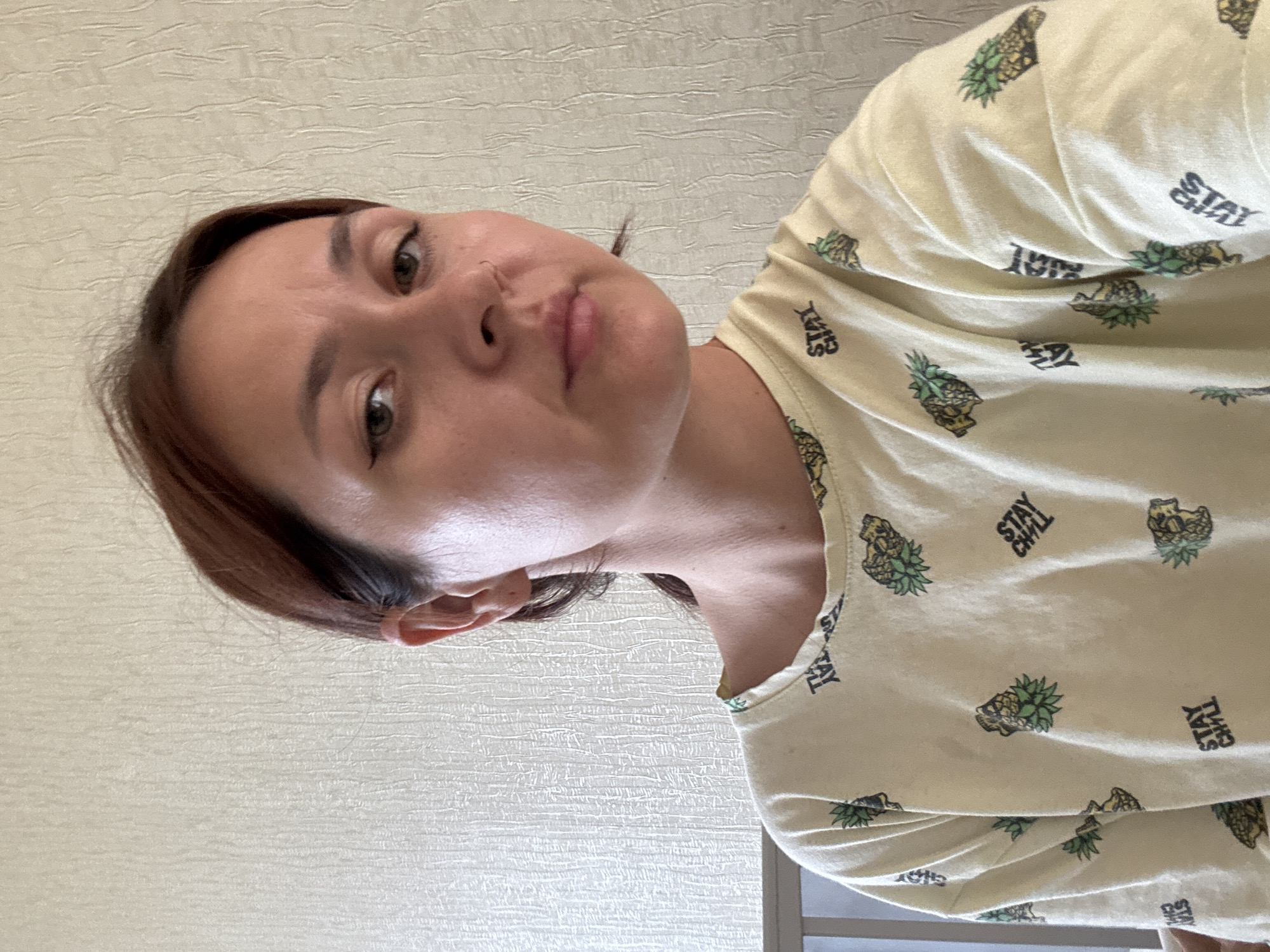 A young woman with brown hair and hazel eyes is looking at the camera. She is wearing a light yellow t-shirt with a pattern of pineapples and the words "STAY CHILL" printed on it. The background is a textured, light-colored wall. The image appears to be a selfie taken indoors, likely in a residential setting in Naberezhnye Chelny, Russia.