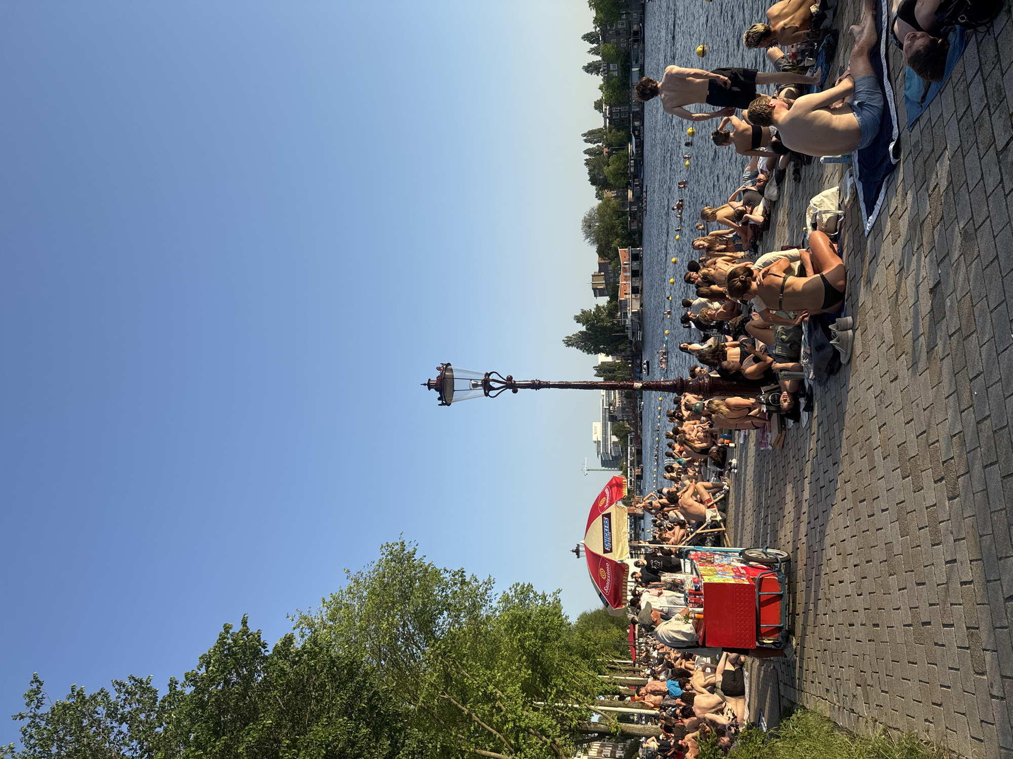 The image depicts a sunny day in Amsterdam, Netherlands, likely during the summer. Many people are sunbathing and swimming along a canal, suggested by the lined yellow buoys. Individuals are sitting or lying on the stone pavement next to the water, some in swimwear. A street lamp and trees line the pavement. A red food or drink cart stands on the walkway, likely selling refreshments to the crowd. Buildings and trees appear on the opposite bank of the canal. The sky is clear and blue.