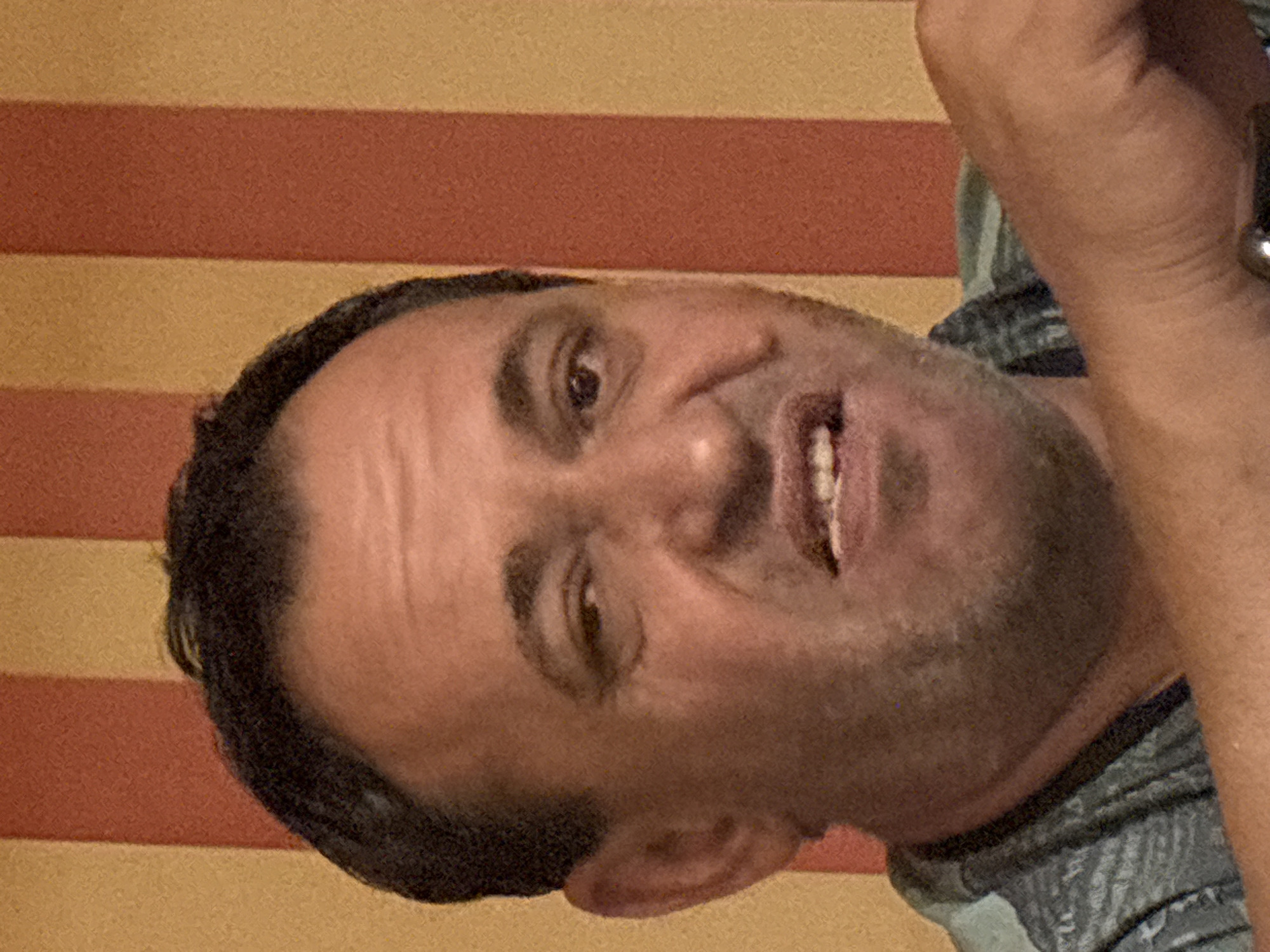 The media file shows a close-up, rotated, and slightly blurry image of a man's face. He appears to be looking towards the camera with his mouth slightly open, revealing some teeth. The man has dark hair and is wearing a grey garment with a striped pattern. The background consists of horizontal stripes of yellow and reddish-brown. There are no discernible location cues, time of day, weather, or visible text. The man's expression could be interpreted as surprised or amused.