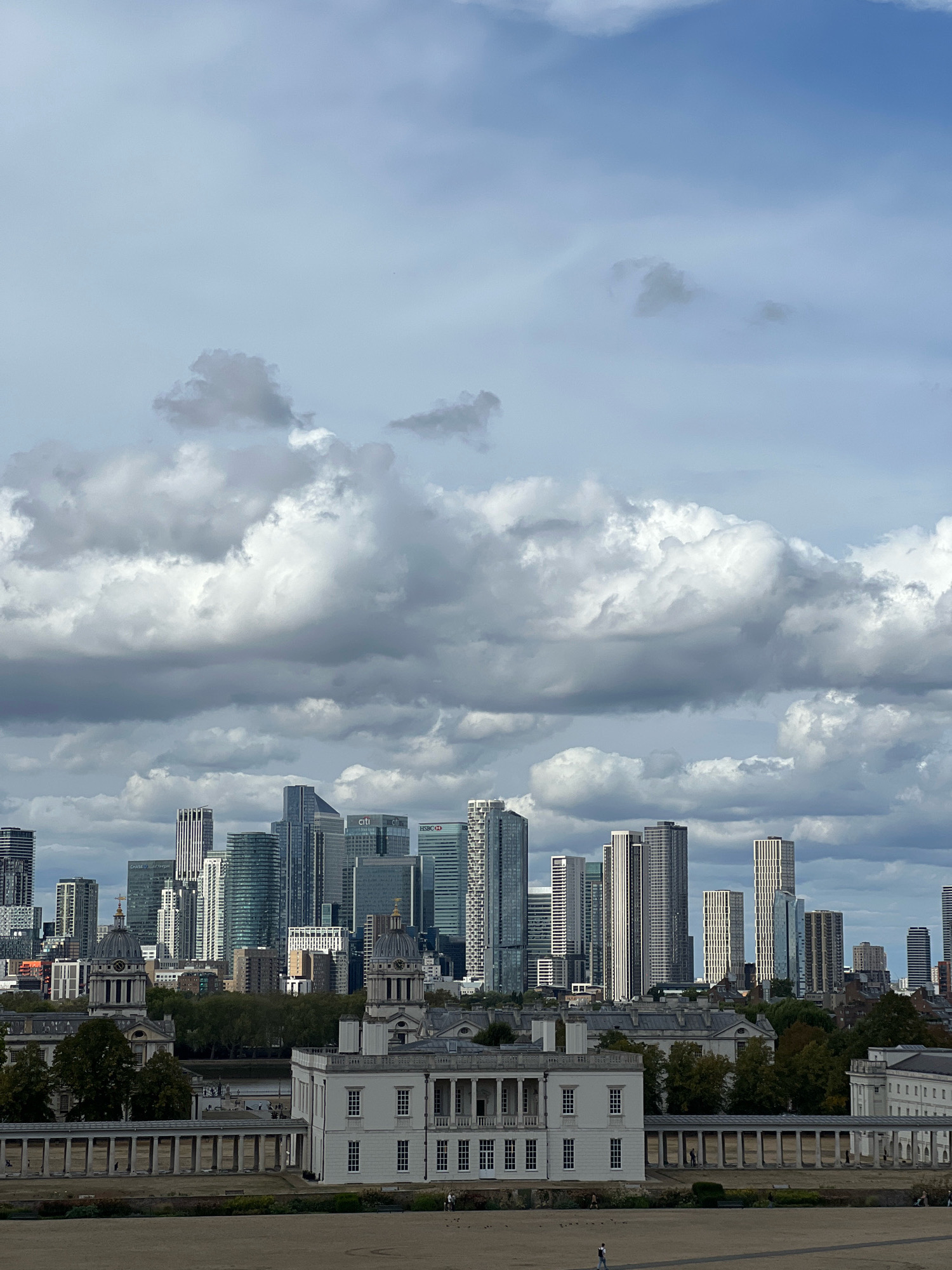 This is a medium-distance, eye-level, vertical, outdoor, daylight shot of a cityscape in London, United Kingdom, featuring a modern business district in the background and historical buildings in the foreground. The sky is overcast with numerous grey and white clouds.
In the background, a dense cluster of modern skyscrapers, characteristic of Canary Wharf, dominate the skyline. These buildings vary in height and architectural style, with some featuring reflective glass facades and others with more geometric, patterned exteriors. Visible signage on some buildings includes "CITI" and "HSBC".
In the middle ground, a strip of green trees separates the historical buildings from the modern skyscrapers.
In the foreground, two large, white, classical-style buildings with domes and colonnades are prominent. These are likely part of the Old Royal Naval College in Greenwich. The building on the right is closer and more detailed, showing rows of windows and a long, covered walkway with columns. The building on the left appears to be a mirror image of the right-side building. Between the buildings, there are pathways and some scattered trees.
In the far foreground, there is a large, open, flat, sandy area, possibly a park or promenade. A few individuals are visible walking on this area. One person is walking on the sandy area towards the right, and another is walking on a path in front of the colonnade of the historical buildings. The overall impression is a juxtaposition of historical and modern architecture under a cloudy sky.