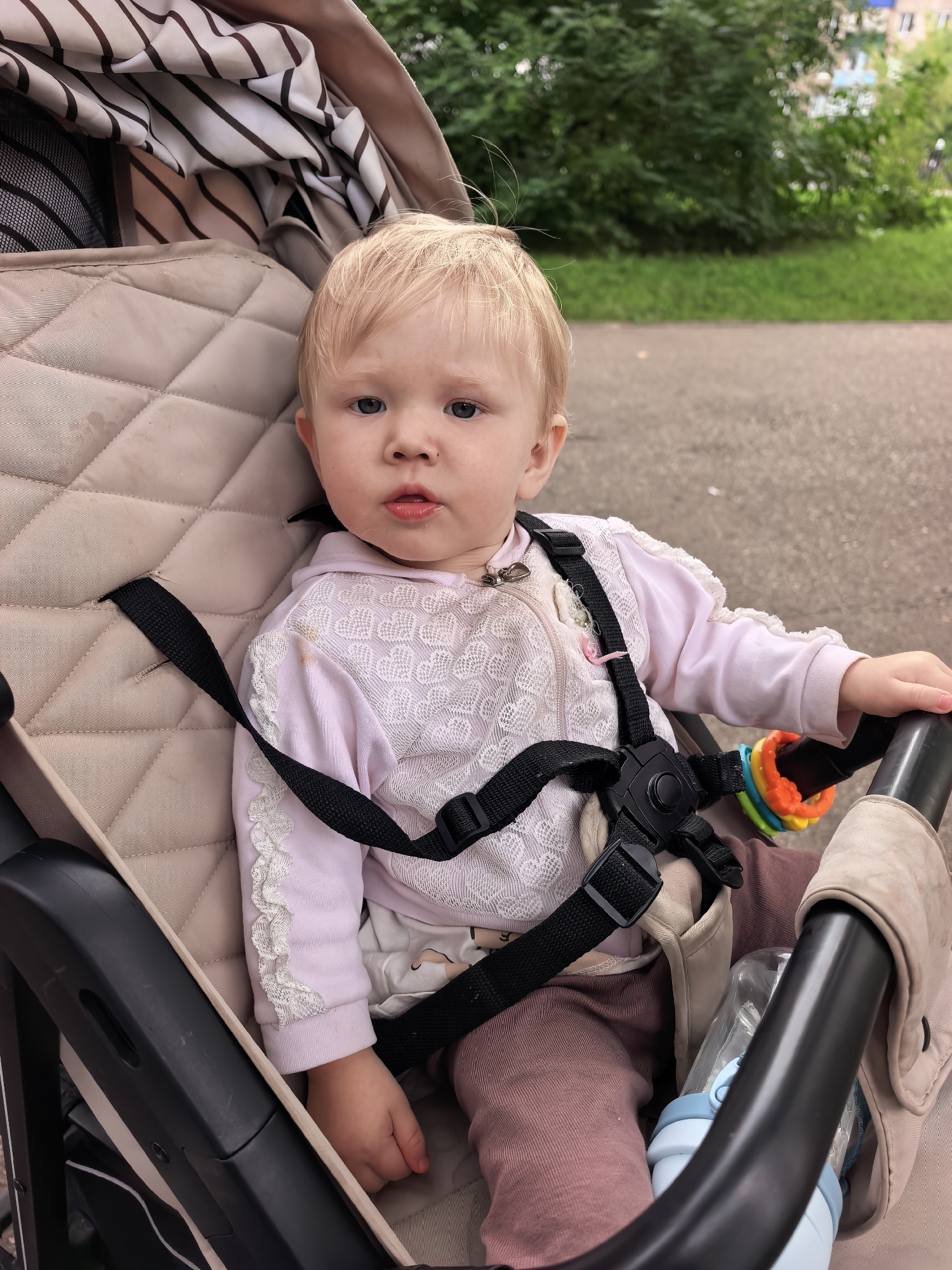 A young child with blonde hair and blue eyes sits in a stroller. The child is wearing a light pink long-sleeved shirt with white lace trim and brown pants. A black harness is secured around the child. A colorful teething toy is attached to the stroller's handle. A water bottle is visible next to the child. The stroller is parked outdoors on a paved surface, with a grassy area and trees in the background. Buildings are visible in the distance. The scene appears to be in a residential area.