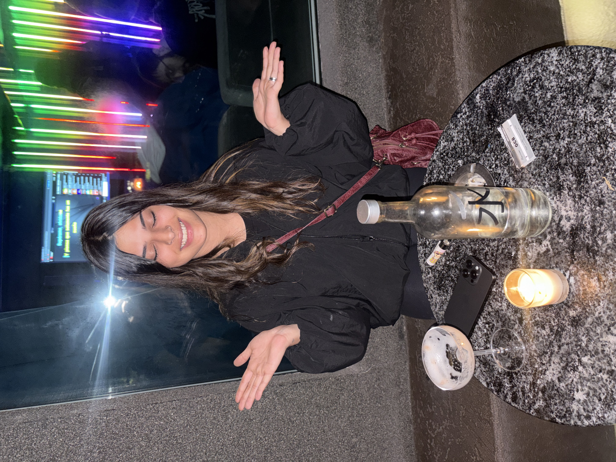 A young woman with long, wavy dark hair, wearing a black jacket, smiles as she sits at a table. She has her hands raised, palms facing outwards. On the table, which has a black and white speckled surface, are a nearly empty wine glass, a black smartphone, a lit candle in a glass container, a nearly empty liquor bottle with a label with the number "12" written on it, a lighter, and a package of rolling papers. In the background, a window reflects colorful neon lights, and a monitor displays a user interface. The location is a bar in Buenos Aires, Argentina.