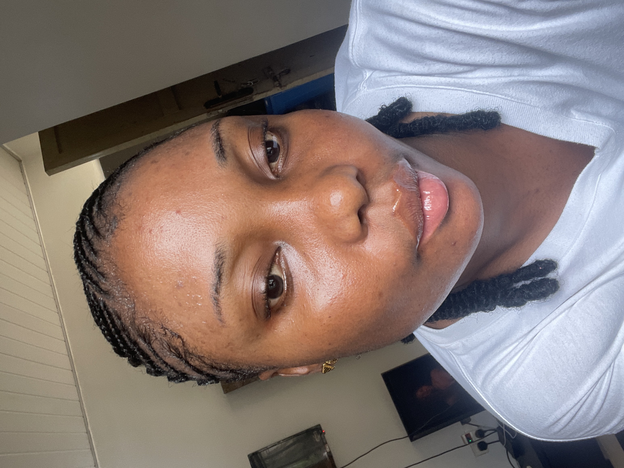 A close-up, angled, indoor portrait of a dark-skinned woman with braids. The woman has dark brown eyes, full lips that appear glossy, and a clear complexion. She is wearing a white t-shirt. Part of a flat-screen TV and electrical outlets are visible in the background. Based on the subject's features and the presence of electrical outlets and a TV in the background, the location is likely a modern residential building in Abuja, Nigeria.