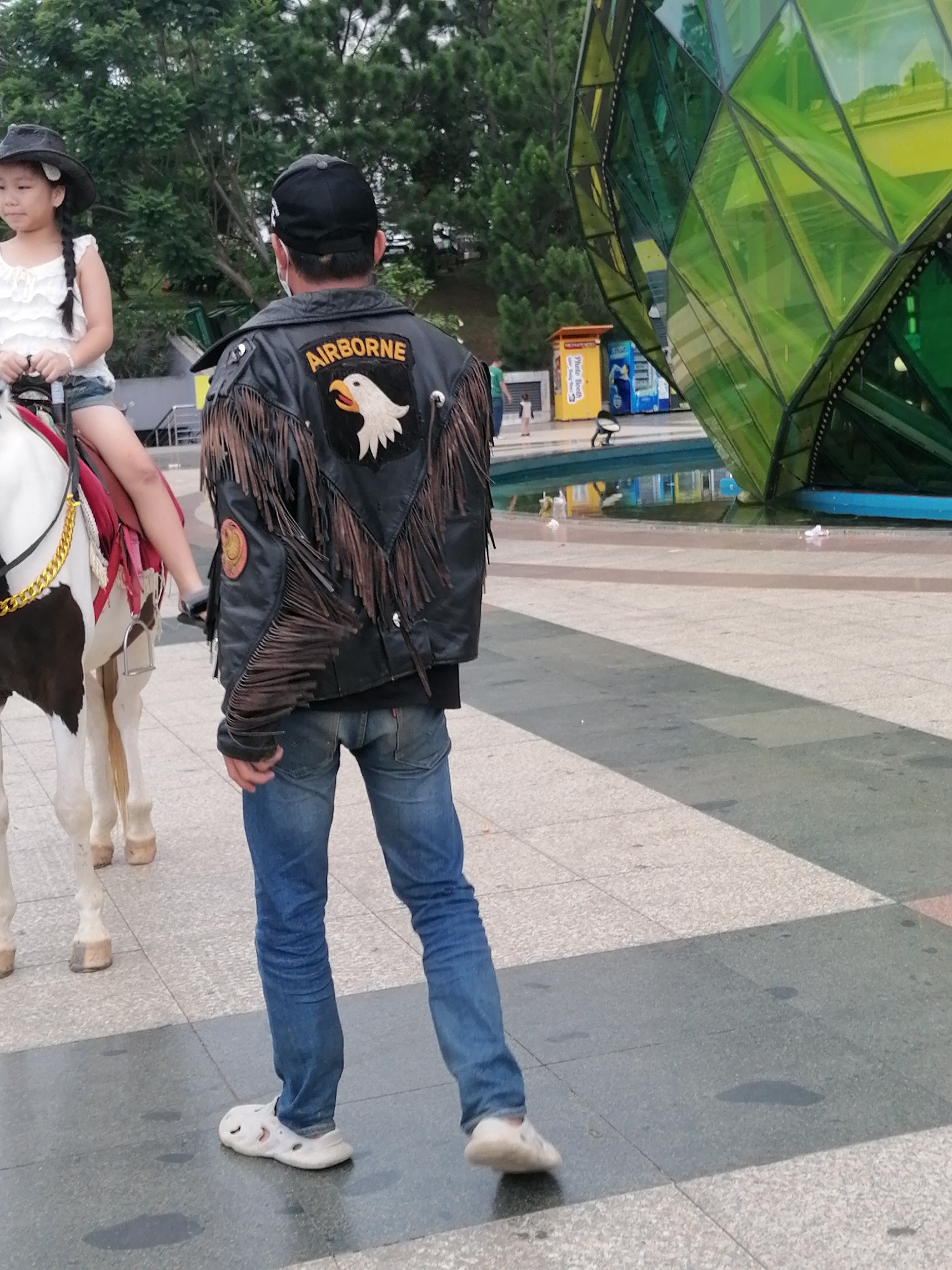 A young East Asian girl with braided hair, wearing a white top and denim shorts, sits on a brown and white pony. She has a dark cowboy hat on her head. Beside her, an East Asian man wearing a black baseball cap, a black leather jacket with fringe and an "AIRBORNE" patch featuring an eagle, and blue jeans walks. He is wearing white Crocs-style shoes. The scene takes place in a paved public area with a pool of water in the background. A large, faceted green structure is visible on the right side of the image. In the background, there are trees, a yellow and blue vending machine, and a small white building labeled "Photo Booth." The overall setting appears to be a park or recreational area in Da Lat, Vietnam.