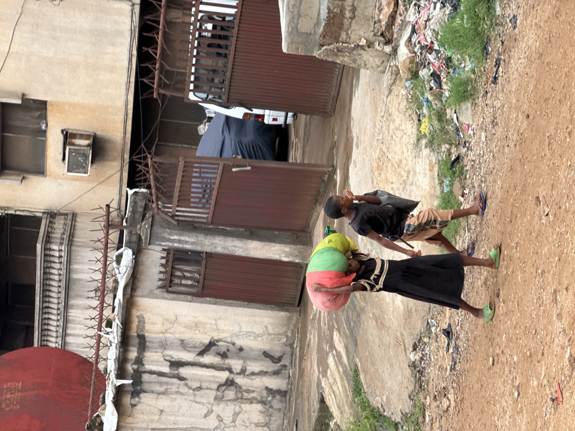 In Onitsha, Nigeria, a child in a black top and patterned shorts walks beside a girl in a black dress who carries a large, multi-colored bundle on her head. Both wear flip-flops. They traverse a dirt path littered with debris and trash, near a building with barred windows, and corrugated metal gates, one open. A heap of garbage with some vegetation sits to the right. The buildings exhibit deteriorated surfaces. A white vehicle is visible behind the gates.
