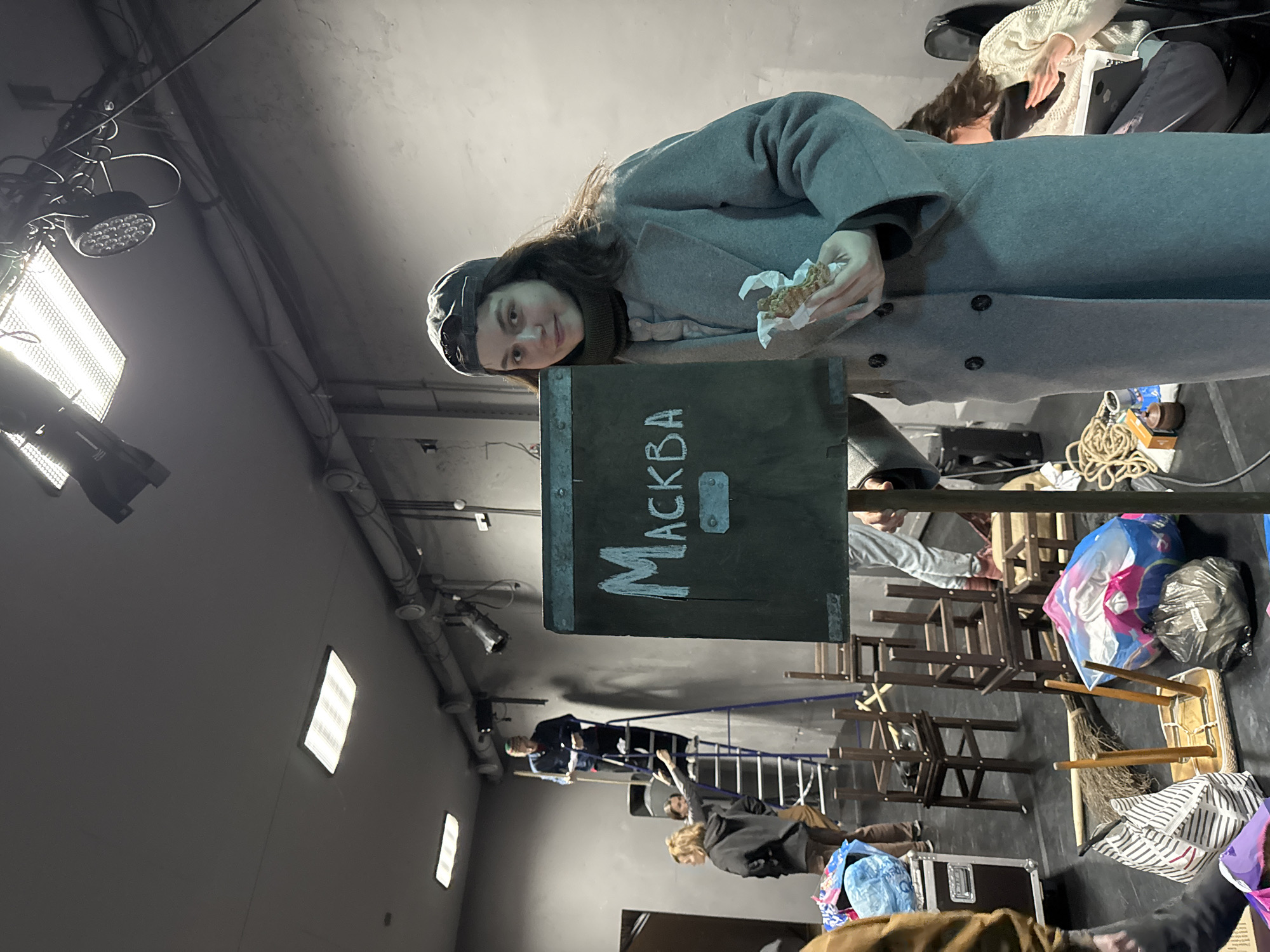 The image shows a person, likely a woman, in a gray coat and cap, holding food in a wrapper. She is standing next to a dark sign with the inscription "MOCKBA" (Moscow in Russian). The location is potentially in a large indoor space resembling a studio or warehouse, with industrial lighting and equipment visible in the background. A ladder, various chairs, and bags filled with objects are also in the shot, suggesting the space may be used for stage production or other work. Several people appear to be working in the distance, walking behind the standing woman, but they are hard to distinguish. Overall, the setting gives off a backstage or industrial ambiance.