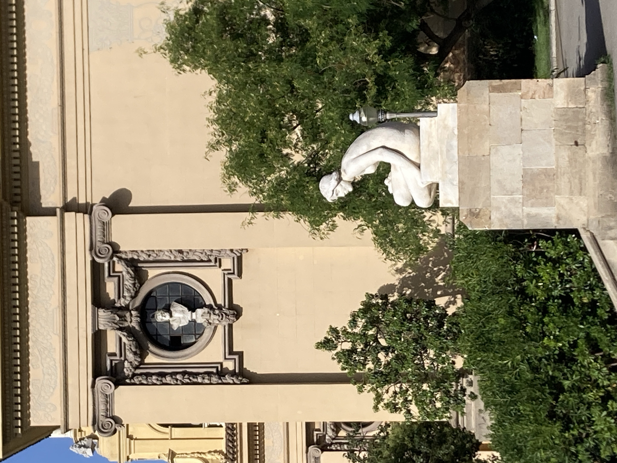 The image shows a sculpture of a seated nude female figure with her head bowed, located in Barcelona, Spain. The sculpture is made of white stone and rests on a stone pedestal. Behind the sculpture, there is a large building with a cream-colored facade, featuring classical architectural elements like columns and a round window with a bust inside. Lush green trees and bushes surround the sculpture and the building, indicating an outdoor setting, likely a park or garden area within the city. The lighting suggests it is daytime, with shadows cast by the trees. The sculpture's style appears to be classical or neoclassical.