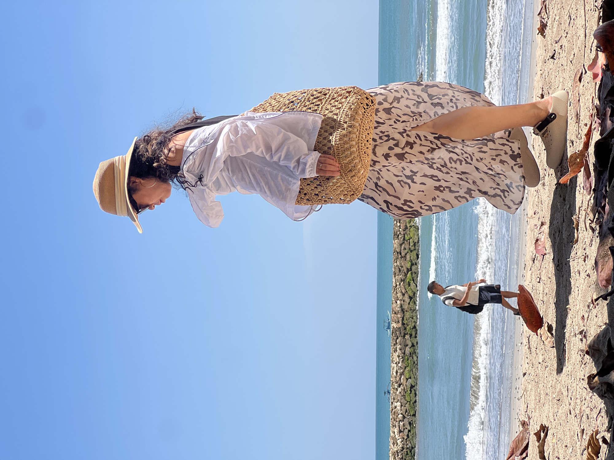 In Kuta, Indonesia, a woman wearing a wide-brimmed hat, sunglasses, a white off-the-shoulder top, and a long, flowing skirt with a leopard-print pattern stands on a sandy beach. She carries a large woven tote bag over her shoulder. Her feet are clad in beige slip-on shoes. In the background, a man with a backpack walks along the shoreline, with gentle waves lapping the beach and a stone jetty with two small boats visible in the turquoise ocean under a clear blue sky. The foreground of the beach is scattered with dried leaves.
