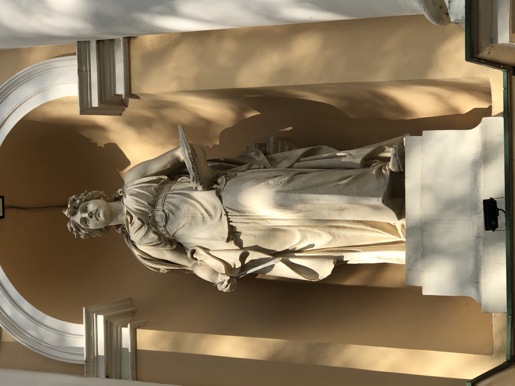 The image depicts a full-body statue of a woman in classical attire, made of light-colored stone, positioned within a recessed architectural alcove. The statue is detailed, with folds in her garment, a laurel wreath on her head, and she holds a tablet or book in her left arm and a stylus or quill in her right hand. The alcove features a curved arch at the top and fluted architectural elements on either side. The background wall of the alcove is a warm ochre color, and the statue is illuminated by natural sunlight, casting shadows on the wall and the statue itself. The statue stands on a multi-tiered white pedestal, which is part of a larger building structure that includes white columns. In the lower foreground, a small black spotlight is visible, mounted on a dark green ledge. The statue is located in Lviv, Ukraine.
