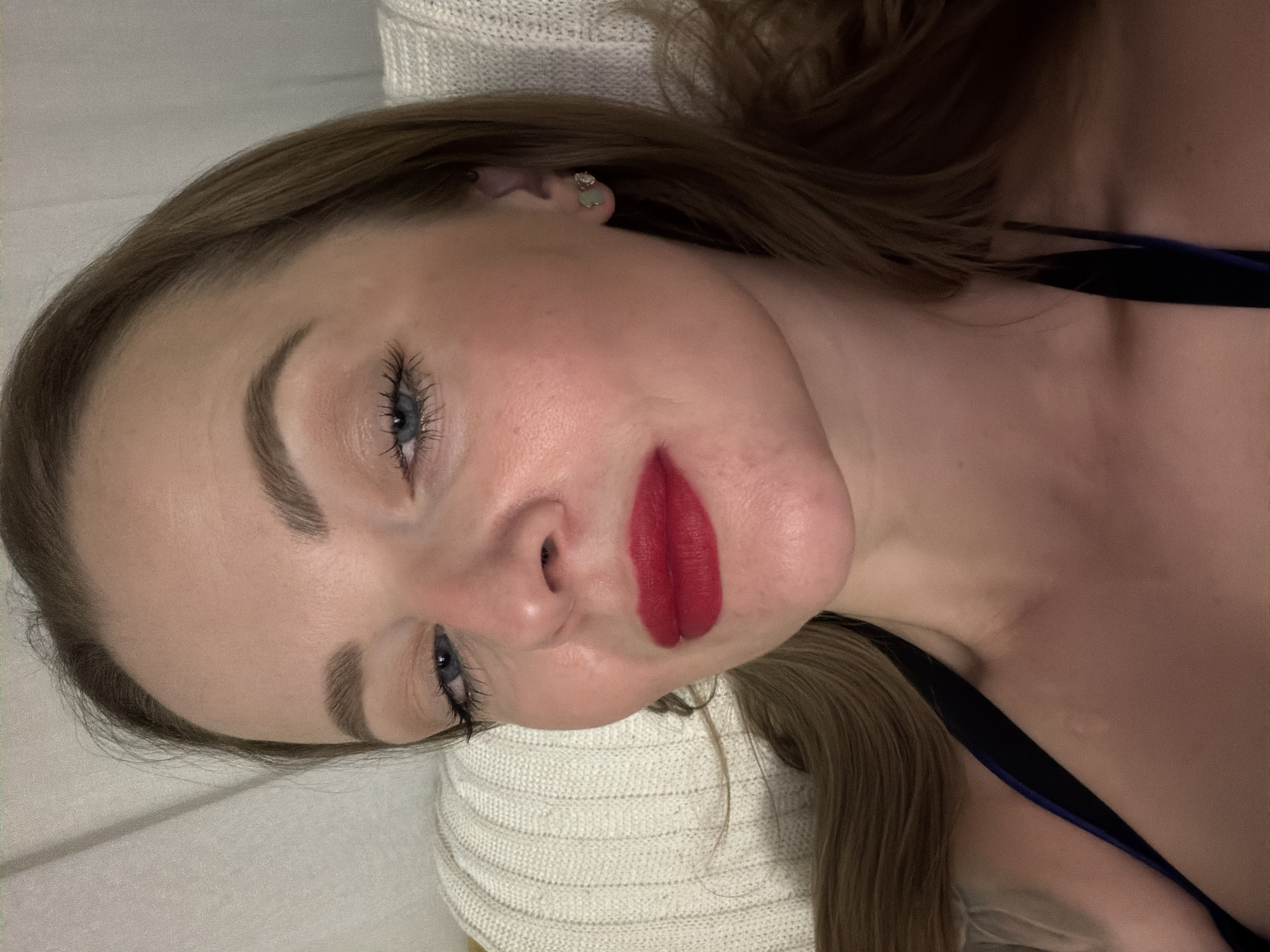 A young woman with light brown hair, blue eyes, and red lipstick is reclining against a white knit surface. She has fair skin and wears an earring in her left ear. The image appears to be taken indoors under warm lighting. A dark strap of clothing is visible at her shoulder. The location in Moscow, Russia is unknown and cannot be determined from the visual information provided.