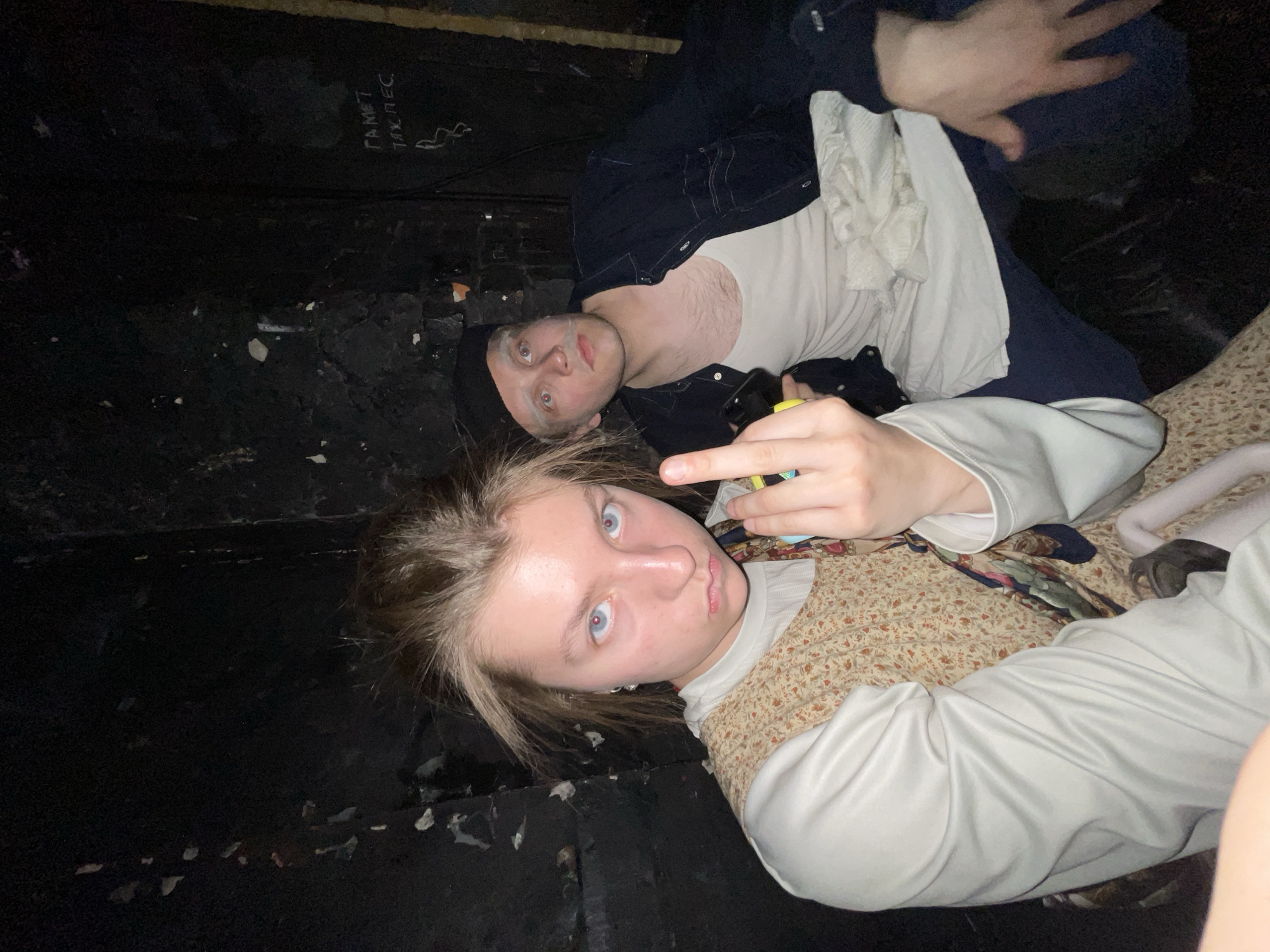 Two individuals are in a dimly lit interior space. One person, wearing a white top and patterned vest, is closest to the camera and gives the middle finger. The other person, wearing a black beanie, white tank top under a dark jacket, is behind the first person. Graffiti text is on the wall.