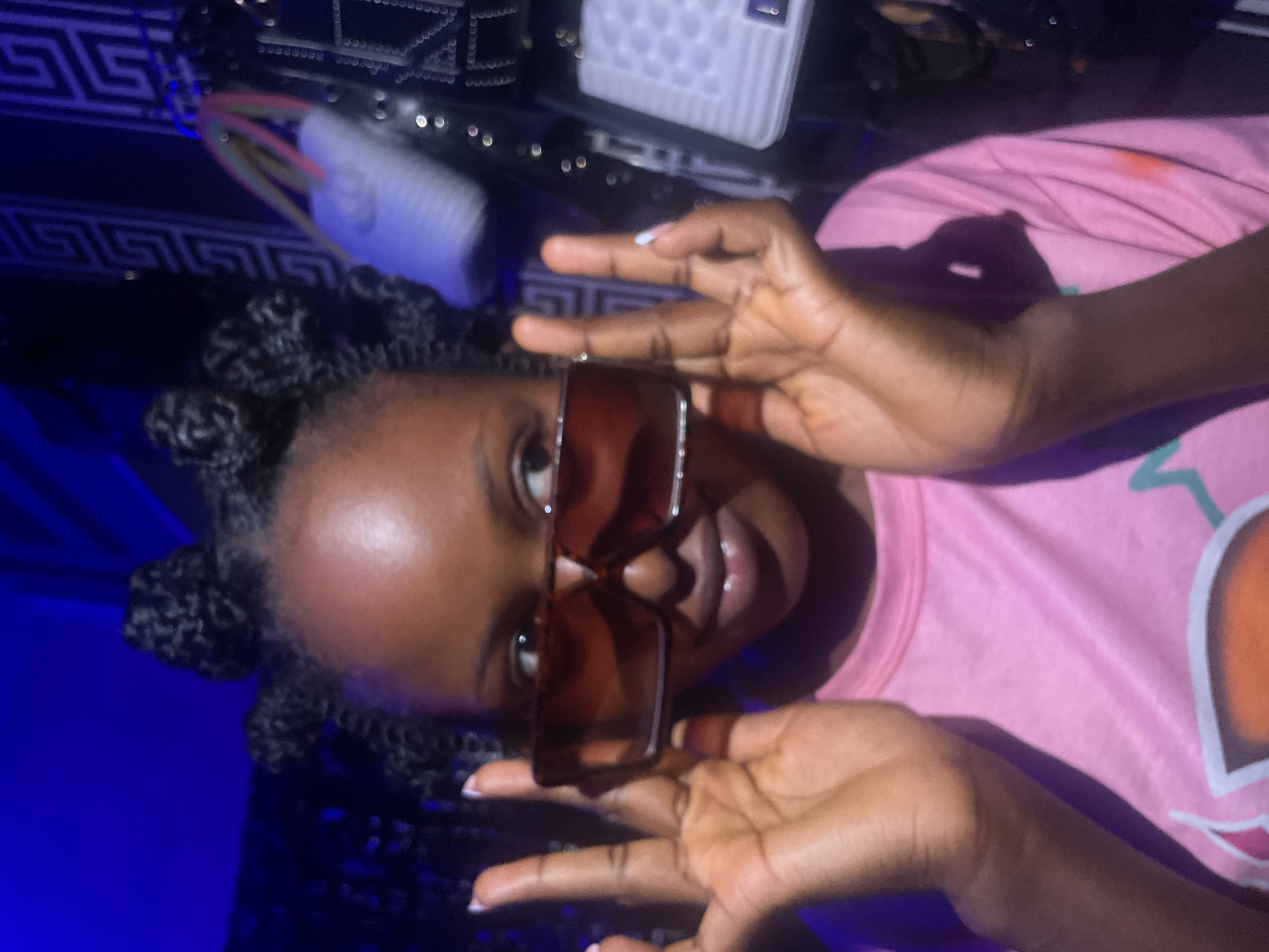 A person with dark skin, wearing a pink t-shirt and brown sunglasses, poses for the camera. Their hair is styled in braided buns. The background is dark with a blue light source and some patterned objects. The location cannot be determined as Lokoja, Nigeria from the image.