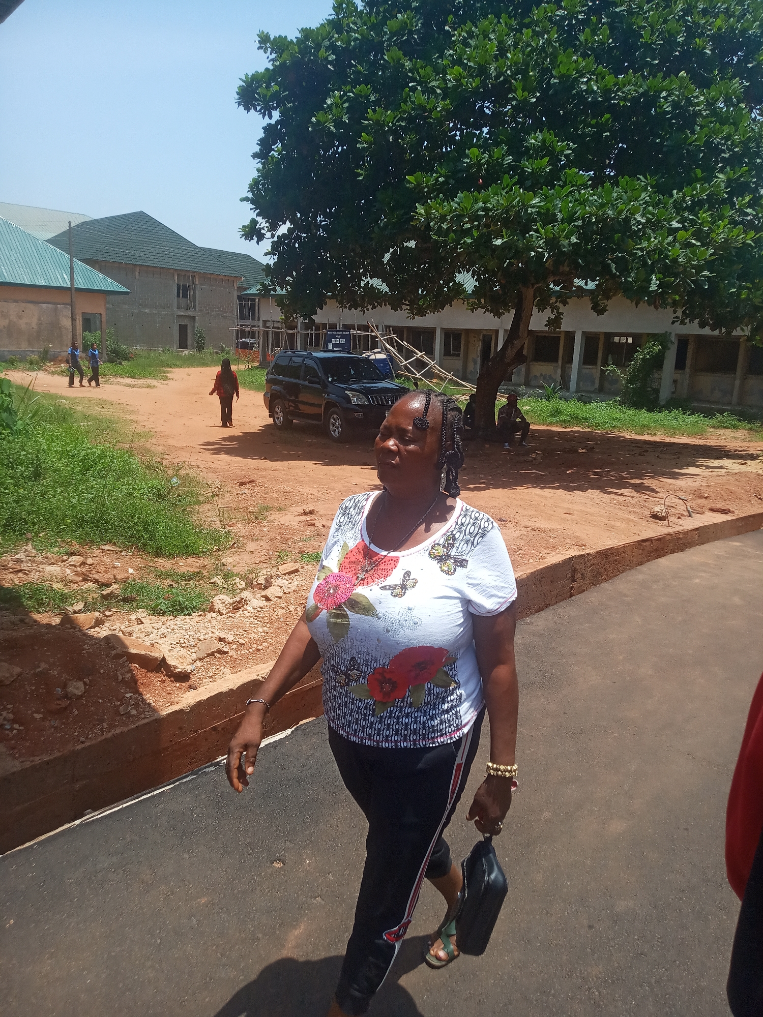 The image shows a woman walking on an asphalt road. She is wearing a white shirt with floral designs, black pants with a red stripe, and sandals. She is carrying a black purse in her left hand and has bracelets on her right wrist.
Behind her, the environment includes a dirt path, a black SUV, green vegetation, and a building with a green roof. In the distance, two people are walking along the dirt path and another person is sitting under a large tree near a building with concrete pillars. The location appears to be Ukwu-Abwa, Nigeria, based on the request.