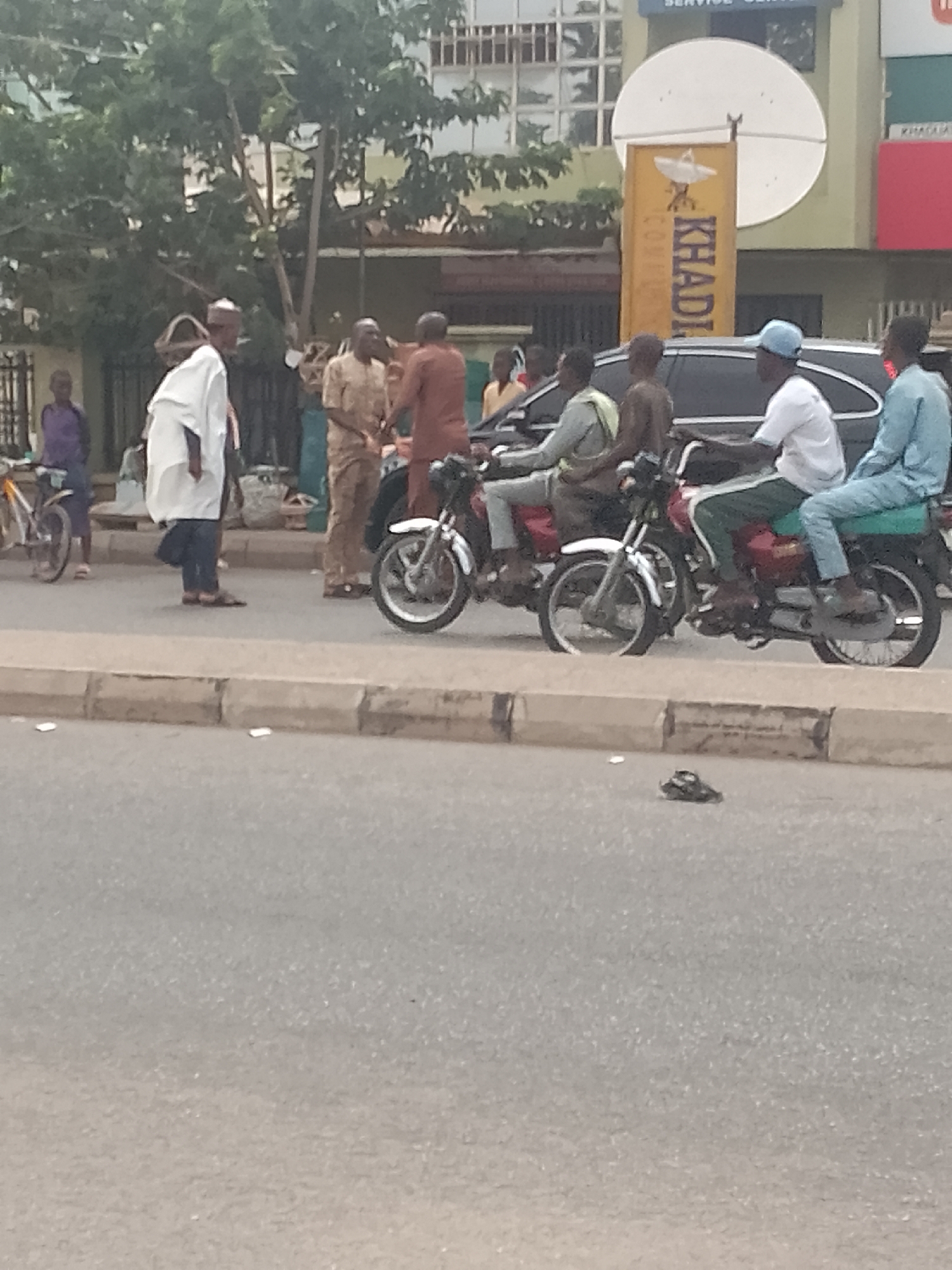 The scene depicts a street in Pantame, Nigeria. There are two motorcycles with riders and passengers on the side of the road. Several people are gathered around near the motorcycles. Buildings, trees, and a parked car form the backdrop of the scene. A shoe lies on the road.
