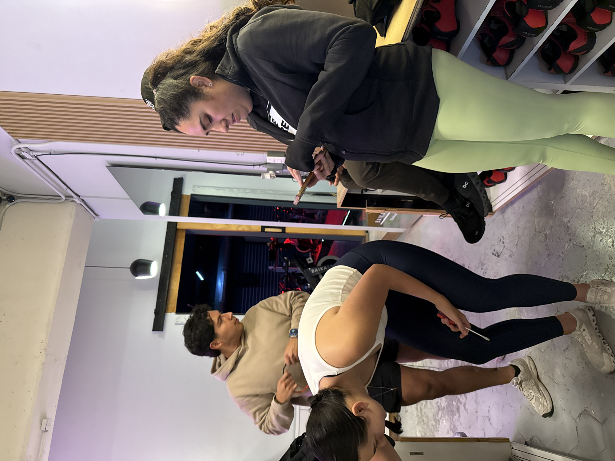 Three people are indoors. One woman with curly dark hair, wearing a black jacket and light green leggings, stands to the left of a dark-haired person in a beige hoodie. The person in the hoodie stands to the left of a dark-haired woman in a white top and dark leggings. Both women hold small objects and are examining the leg of the woman in the white top. Shelves of shoes are to the right. The city of Puebla is not visible in the image.