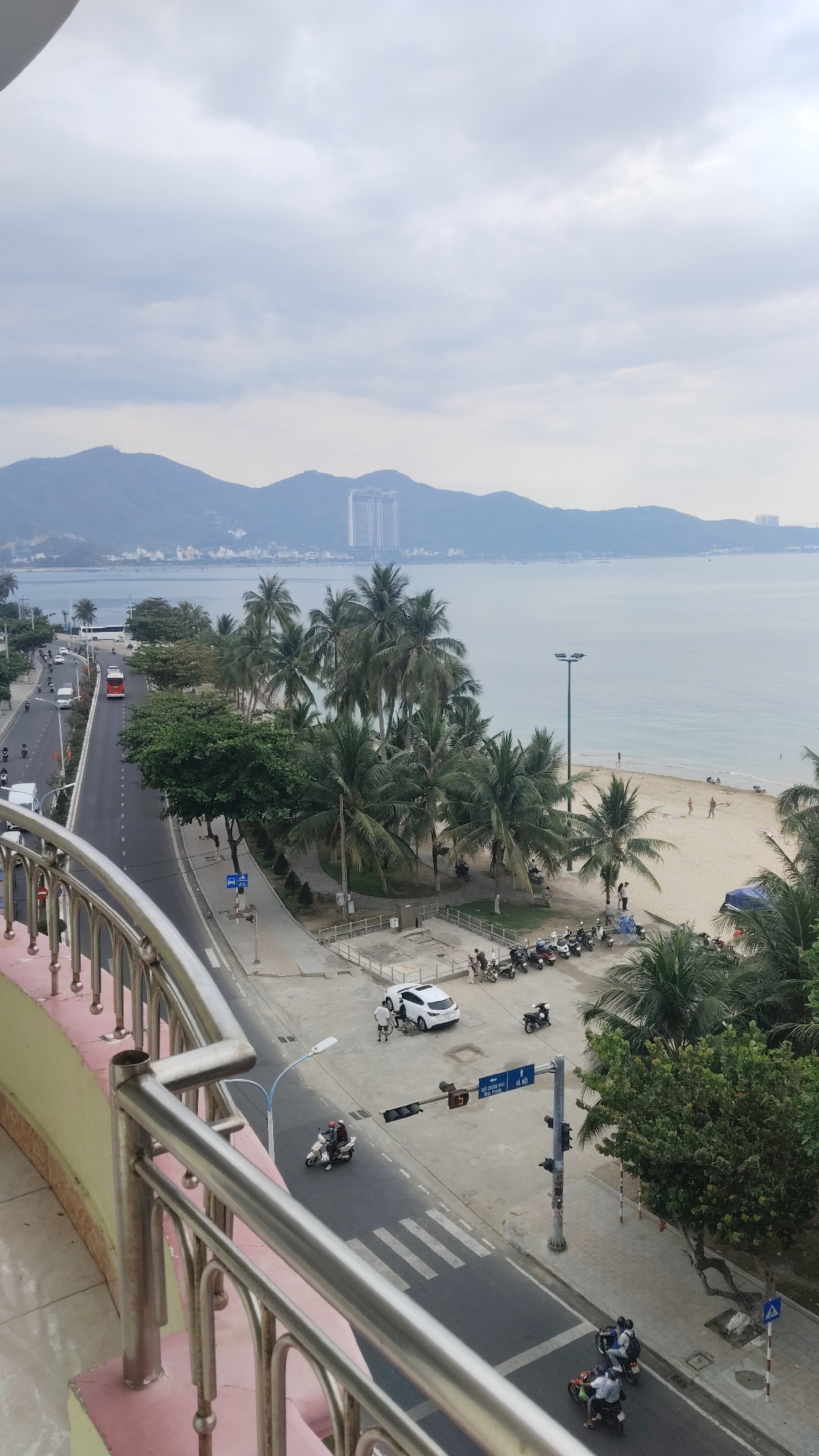 The image presents a high-angle view of a coastal urban scene in Bac Nha Trang Ward, Vietnam. The foreground is dominated by a metallic balcony railing, partially obscuring the lower left corner. A road runs parallel to the coastline, with light vehicular and motorcycle traffic. A zebra crossing is visible at a road intersection, accompanied by a traffic light.
On the right of the road, a row of parked motorcycles are present along the pavement, next to a sandy beach. Palm trees are interspersed between the road and the beach, providing shade. People are sparsely scattered on the beach, some appear to be walking or relaxing.
In the background, there is a coastline and a body of water, possibly the sea or a bay. Further in the distance, a mountain range is visible under an overcast sky. A tall building stands out against the mountain backdrop.