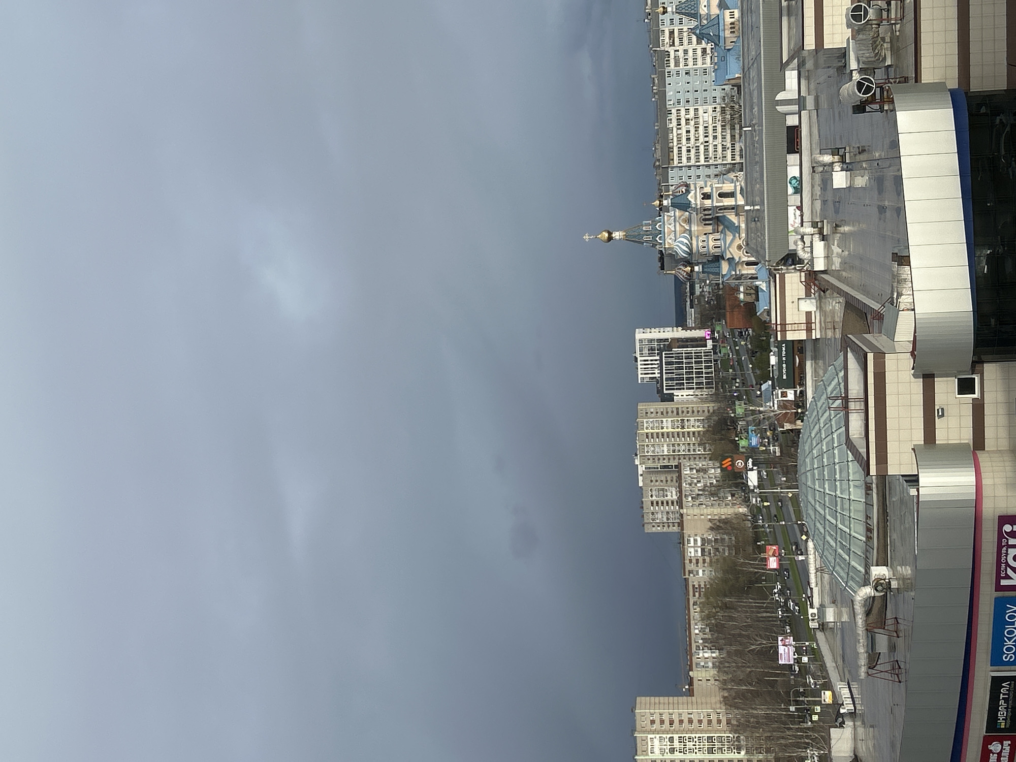 The media file presents a high-angle view of a cityscape in Izhevsk, Russia, under an overcast sky. The dark, cloudy expanse dominates the upper and left portions of the image, suggesting gloomy weather or impending rain.
In the mid-ground and foreground, a collection of urban structures is visible. On the right, a large commercial building, possibly a shopping center, features a modern facade with white and blue panels and a glass-domed section on its roof. Visible on this building are ventilation units and signage.
Further into the city, a distinctive church with traditional Orthodox architecture stands out. It boasts multiple golden onion domes and blue roofs with decorative patterns, serving as a prominent landmark. Surrounding the church and extending into the background are numerous multi-story residential and commercial buildings, forming a dense urban fabric. Distant roads, scattered trees, and other urban infrastructure are also discernible.
Several signs with Cyrillic text are visible on the buildings, including:
* "КВАРТАЛ" (KVARTAL), likely a district or shopping center name.
* "ВКУСНО-И ТОЧКА" (VKUSNO-I TOCHKA), a Russian fast-food chain.
* "SOKOLOV", a jewelry brand.
* "ЕСЛИ ОБУВЬ, ТО" (ESLI OBUV', TO), translating to "If shoes, then..."
* Partial text like "AL", "Илч", and "втория мужского сти".
The scene depicts a typical urban environment during daytime, characterized by the heavy cloud cover that creates a muted lighting effect. No specific human activity is clear due to the distance.