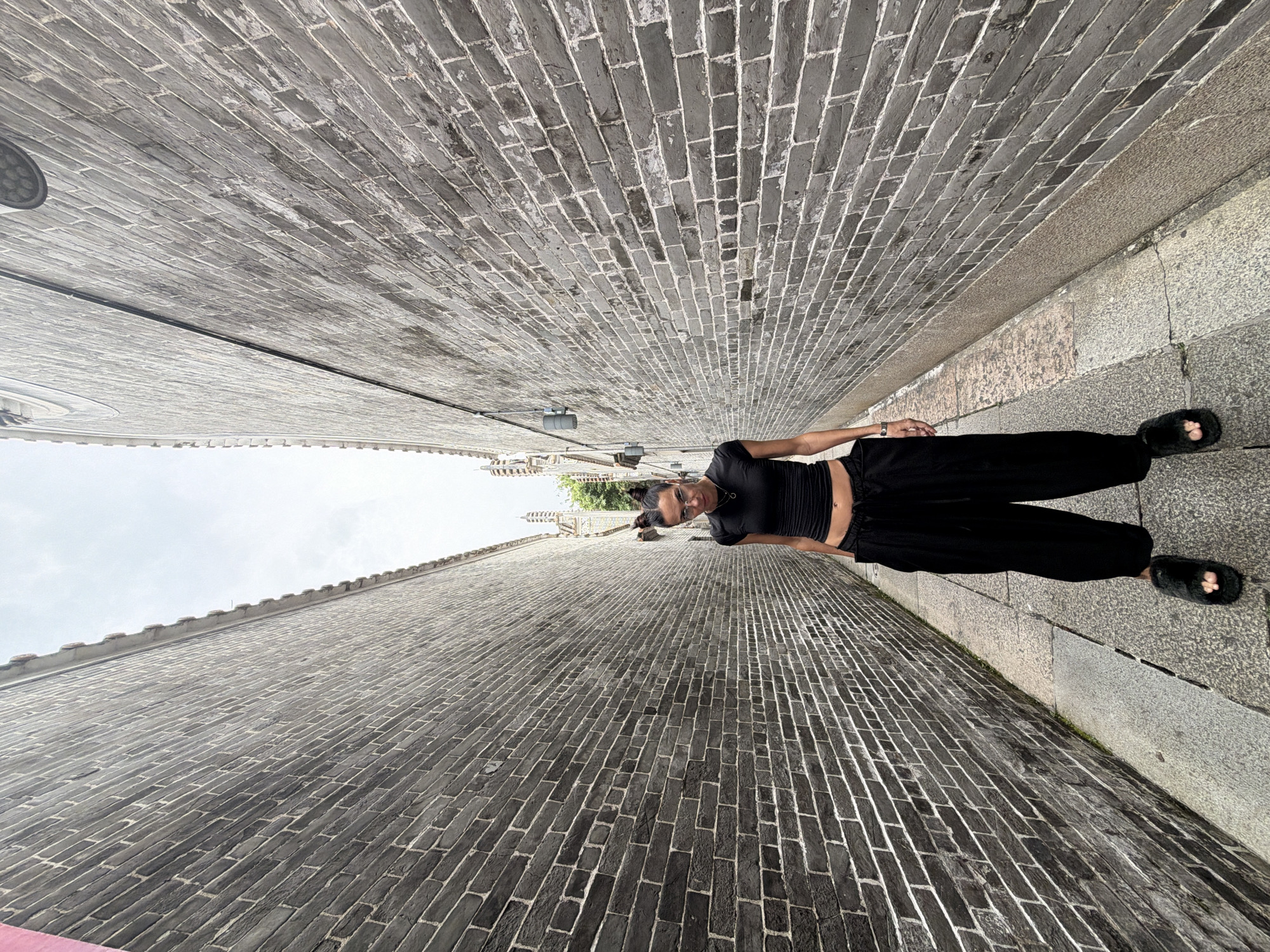 A woman stands in a narrow alleyway in Chancheng District, China. She is facing the camera and is dressed in a black crop top and black wide-leg pants, with fluffy black slippers. The alley walls are made of grey bricks, and the ground is paved with stone. The sky is overcast.