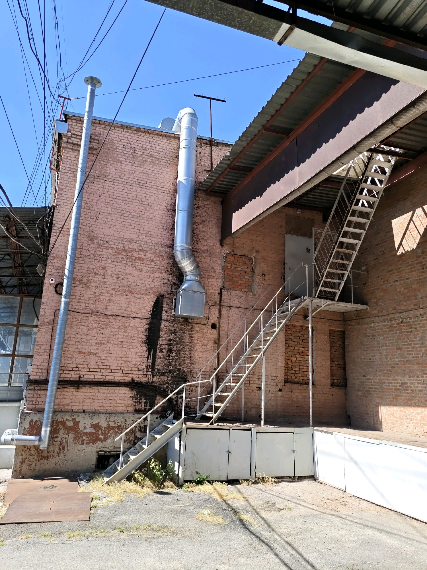 The image depicts an exterior view of a brick building in Rostov-on-Don, Russia. A prominent feature is a long, silver ventilation pipe running vertically along the pinkish-red brick wall. Another, wider ventilation pipe with a curved section is also visible, attached to the upper part of the building. An external metal staircase, painted white, ascends to a platform with a door on the second level. The building has a rough, weathered lower section with peeling paint. Overhead, a corrugated metal roof structure extends from the building, supported by metal beams. A complex network of electrical wires is strung across the clear blue sky. The ground in front of the building is a mix of asphalt and dirt, with some sparse vegetation. Several white storage units with doors are positioned at the base of the building and platform. The overall impression is of an industrial or utility area with signs of wear and tear.