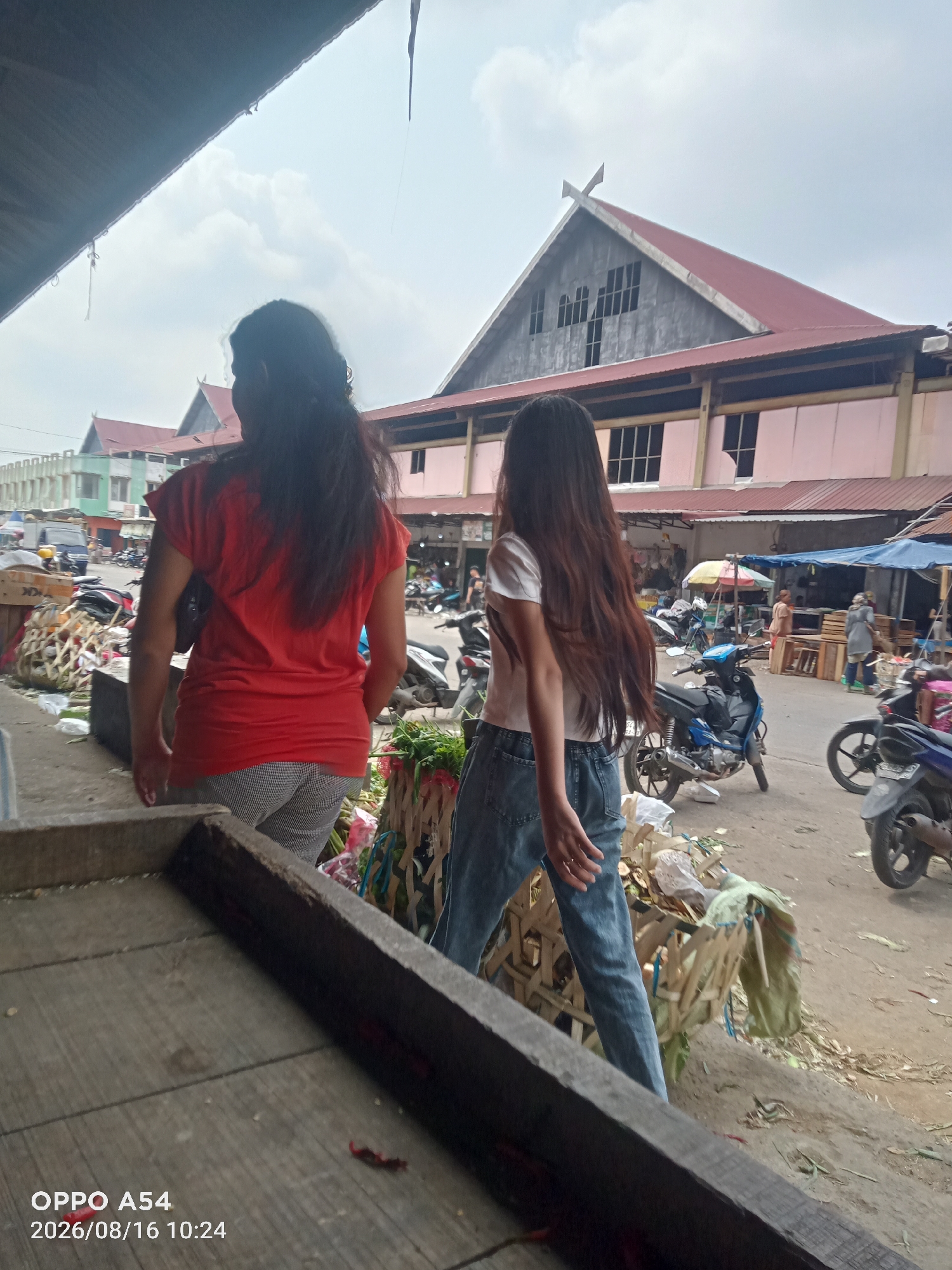 The image depicts a bustling outdoor market scene, likely in Jambi, Indonesia, given the timestamp and context. In the foreground, two young women are the primary subjects. The woman on the left, facing away from the camera, wears a bright red t-shirt and patterned trousers. Her dark, long hair is visible. The woman on the right, also facing away, has on a white t-shirt and blue jeans, with her long brown hair flowing.
The market environment is characterized by a covered structure with a distinctive red-tiled roof and a large, grey, triangular facade with windows, suggesting a substantial building, possibly a marketplace or a large shop. Stalls and displays of goods are visible throughout the scene. In the middle ground, various motorcycles are parked, indicating a common mode of transport. Piles of produce or other goods, some wrapped in plastic and tied with string, are scattered around the market floor. Further back, more market stalls and buildings can be seen, some with blue tarps providing shade. People are present in the background, engaged in activities typical of a market. The ground appears to be unpaved, with debris and vegetation visible. The overall atmosphere is one of a lively, somewhat disorganized, but active marketplace. The image captures a moment of everyday life in what appears to be a typical Southeast Asian market setting.
