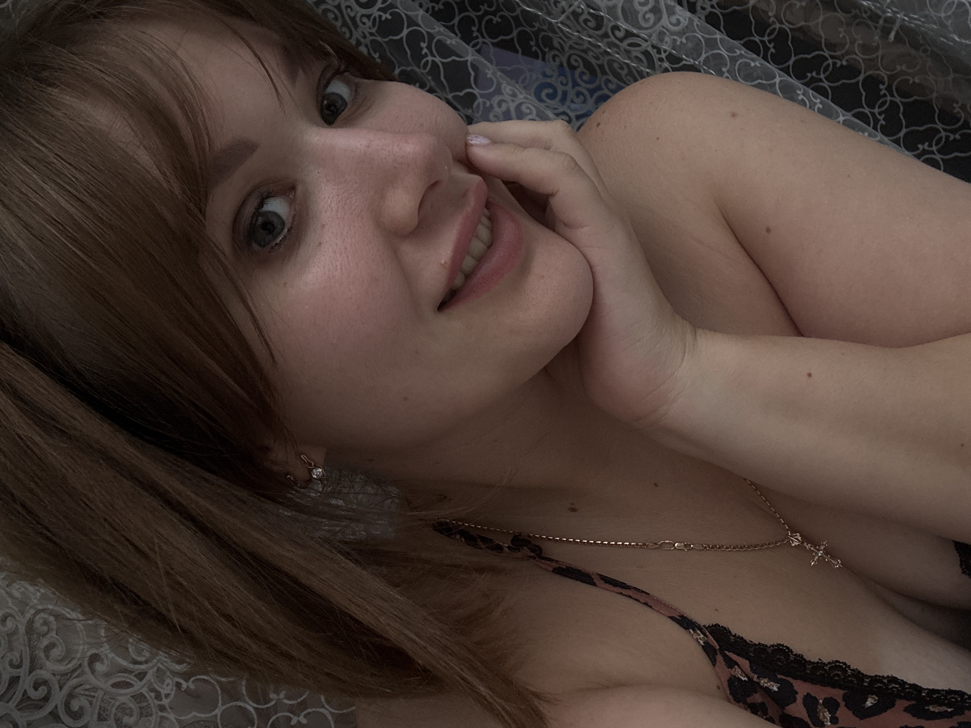 A young woman with auburn hair and light skin is pictured in a close-up, selfie-style shot. She is resting her head on her hand, with her eyes looking towards the camera and a slight smile on her lips. She is wearing a leopard-print top with black lace trim and a delicate gold necklace with a cross pendant. The background appears to be a bed with patterned sheets and a curtain. The image is grainy and dimly lit, with a soft focus. The location of Chaikovsky, Russia is not evident in the image.