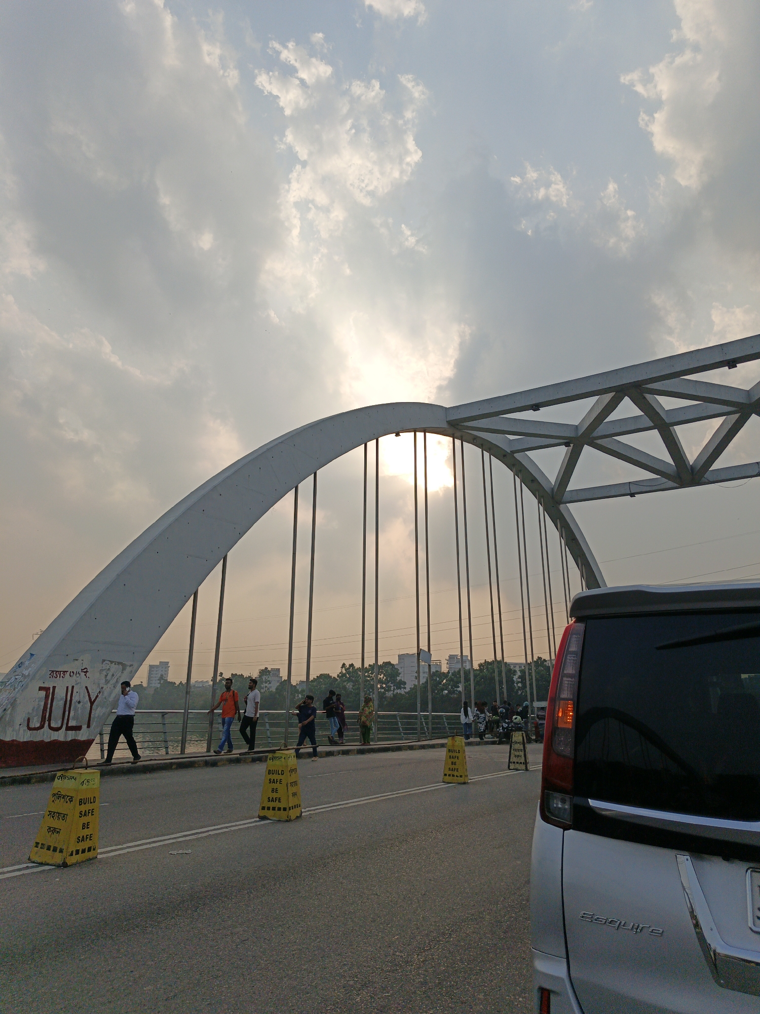 The image depicts a bridge scene in Dhaka, Bangladesh, under a cloudy sky with the sun visible behind the bridge's structure. The bridge has a prominent arch, and the word "JULY" is painted on one side. Several pedestrians are walking along the bridge's pathway. Yellow traffic barriers labeled "BUILD SAFE BE SAFE" in both English and Bengali are placed along the road, presumably to demarcate a lane or construction zone. A silver car, with "Esquire" inscribed on its rear, is partially visible in the foreground on the right side of the frame. In the background, a cityscape is faintly visible. The time of day appears to be late afternoon or early evening, given the soft light and shadows.