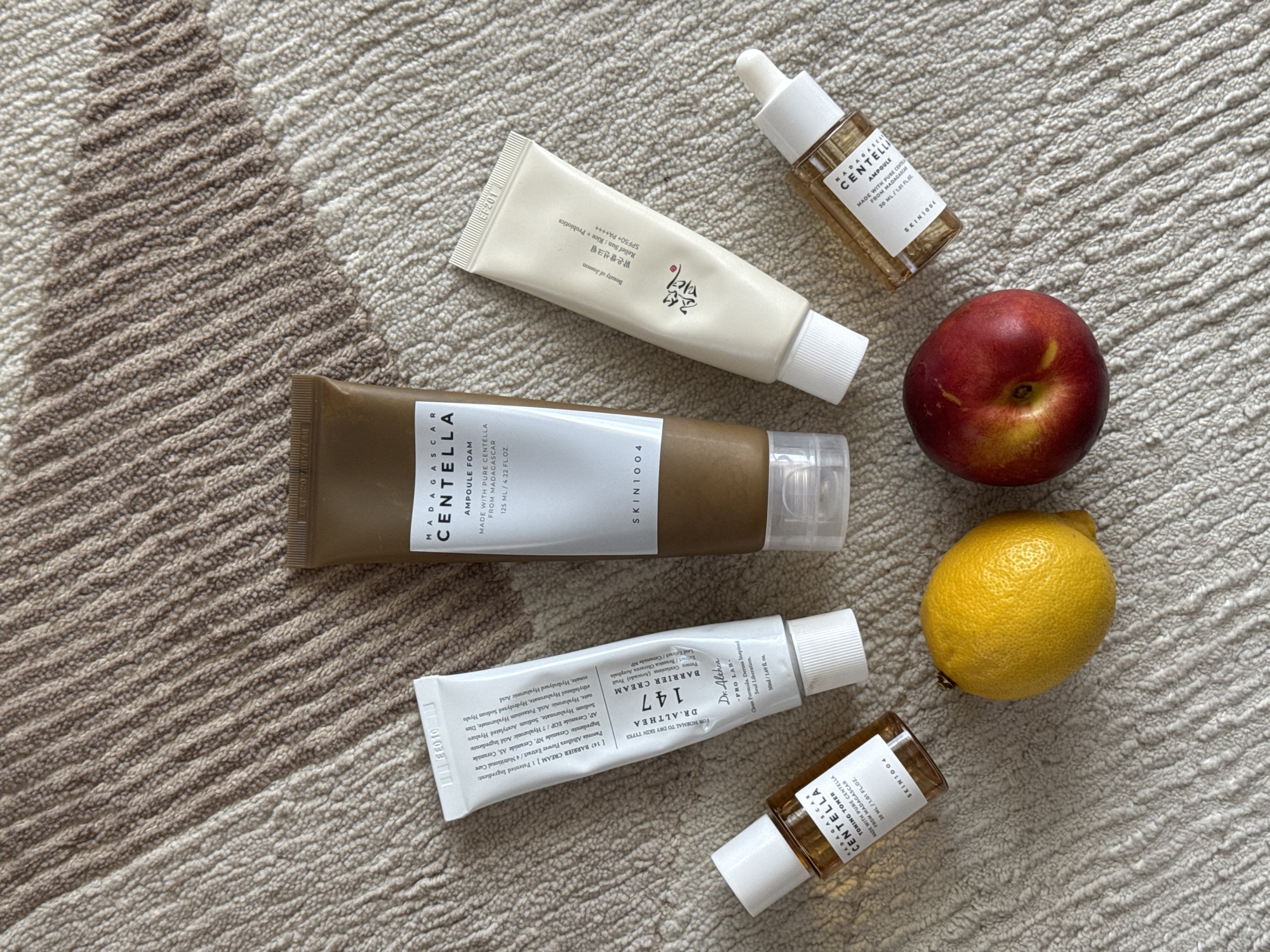 The image shows several skincare products arranged on a textured rug with a beige and brown pattern. There are four tubes and two bottles of skincare products, including brands like SKIN1004 and Dr. Althea. The products include cleansers, serums, and creams. Additionally, a nectarine and a lemon are placed among the skincare items. The environment appears to be an indoor setting. There is no information available about the location of Podgorica, Montenegro in the image. There are no people, actions, or interactions present in the image.