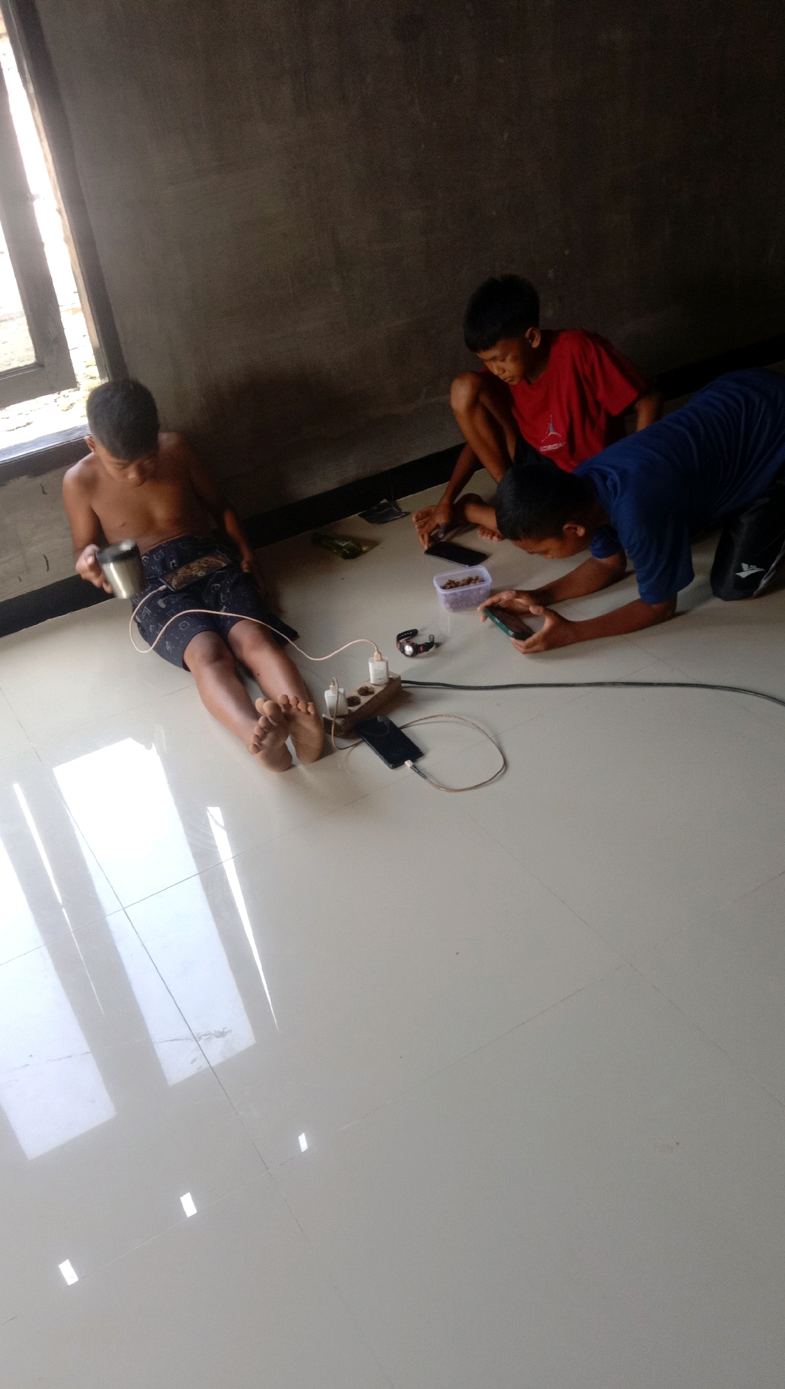 Three young boys are sitting on a light-colored tiled floor in what appears to be an indoor setting. The background is a dark gray wall. To the left, a window with sunlight streaming through is visible. One boy, shirtless and wearing patterned shorts, is leaning back, holding a metal cup in his left hand and looking down at his phone. His phone and a charger are plugged into a power strip. Another boy, wearing a red t-shirt, is crouched in the middle, also looking at a phone. A third boy, wearing a blue t-shirt, is leaning forward on his hands and knees, intensely focused on a phone he holds. A charger is also connected to his phone. In front of the boys, a power strip is connected to multiple devices. A watch is lying near the power strip, and a small plastic container filled with what looks like snacks is also present. The overall scene suggests a casual, relaxed atmosphere, with the boys engrossed in their mobile devices, possibly playing games or watching videos. The lighting indicates it is daytime. The location is identified as Kota Pari, Indonesia. There is no visible text in the image.