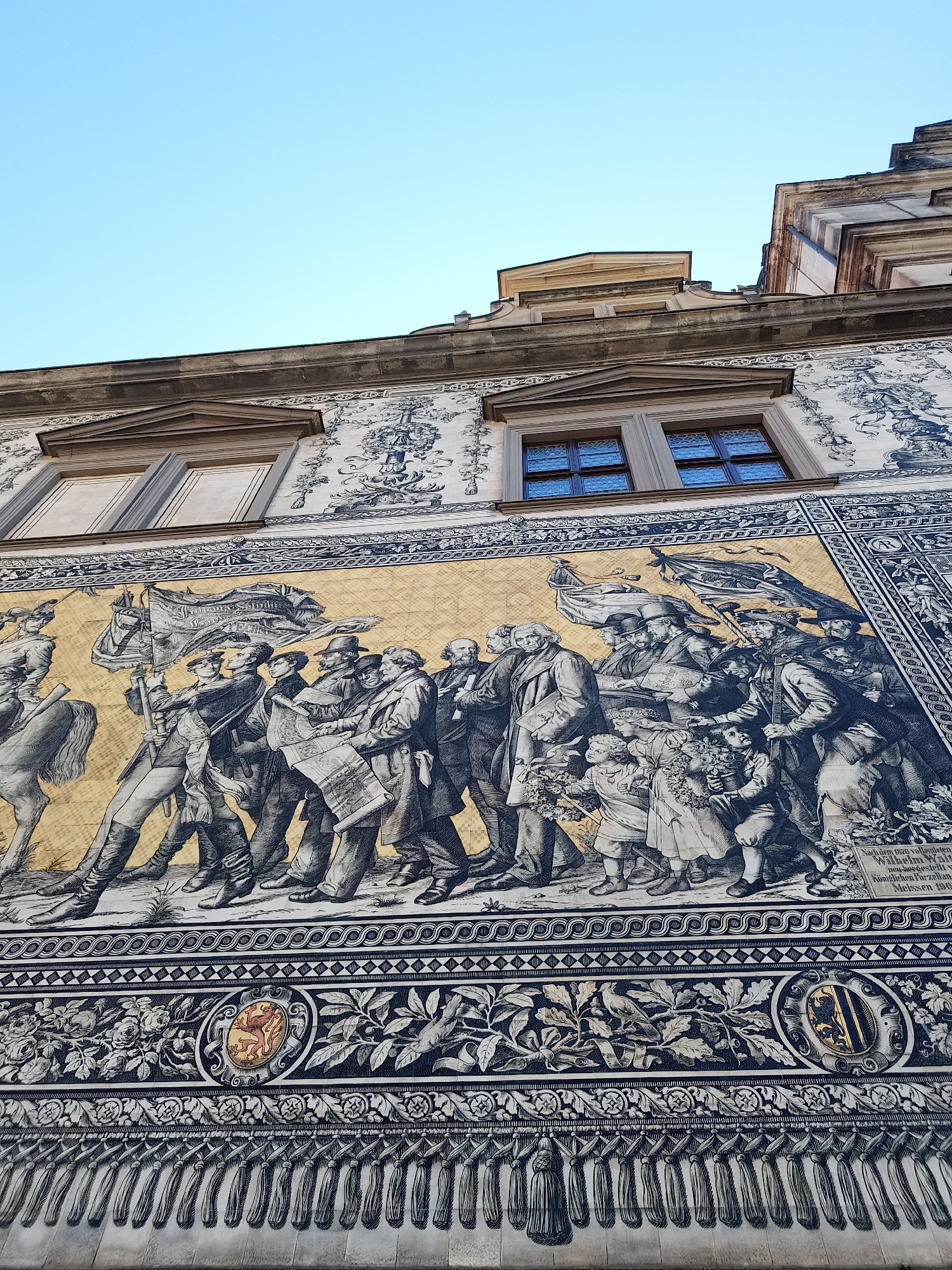 The image shows the Fürstenzug (Procession of Princes) in Dresden, Germany. This is a large porcelain mural on the outer wall of the Stallhof (Stable Yard) of Dresden Castle. The mural depicts a mounted procession of the rulers of Saxony from the 12th to the 19th century. The mural is made of approximately 25,000 Meissen porcelain tiles. The location of this mural is in the city of Dresden, Germany.