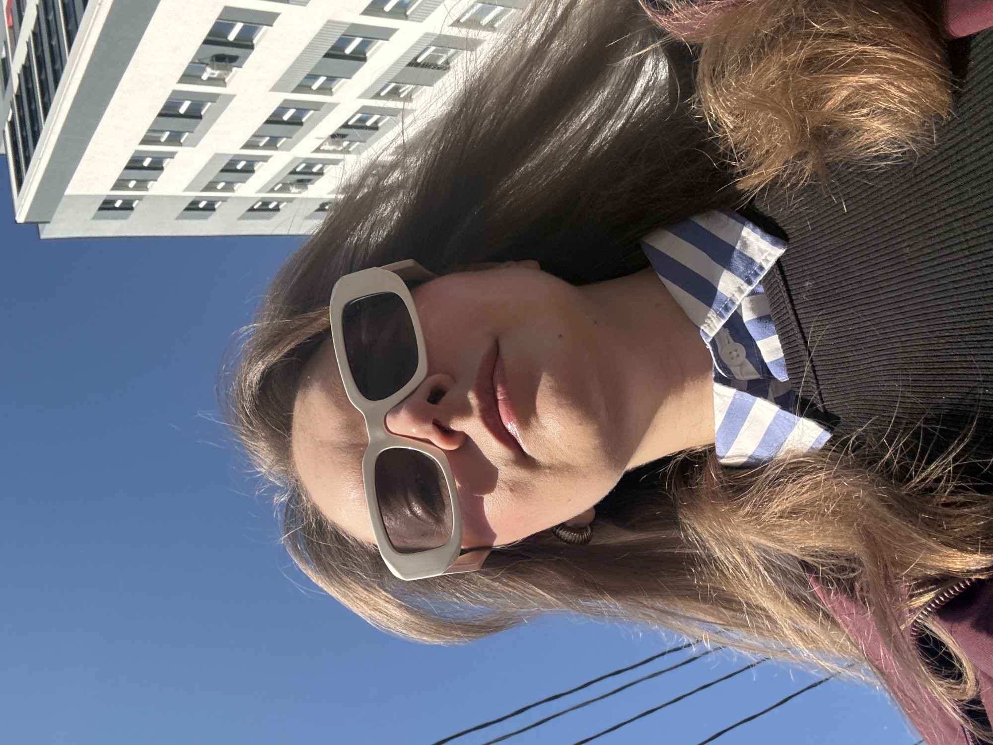 A woman wearing sunglasses, a black ribbed sweater, and a blue and white striped shirt stands in Izhevsk, Russia. A tall white building with multiple rows of windows is behind and above her to the left. Power lines are visible in the foreground. The sky is clear and blue.