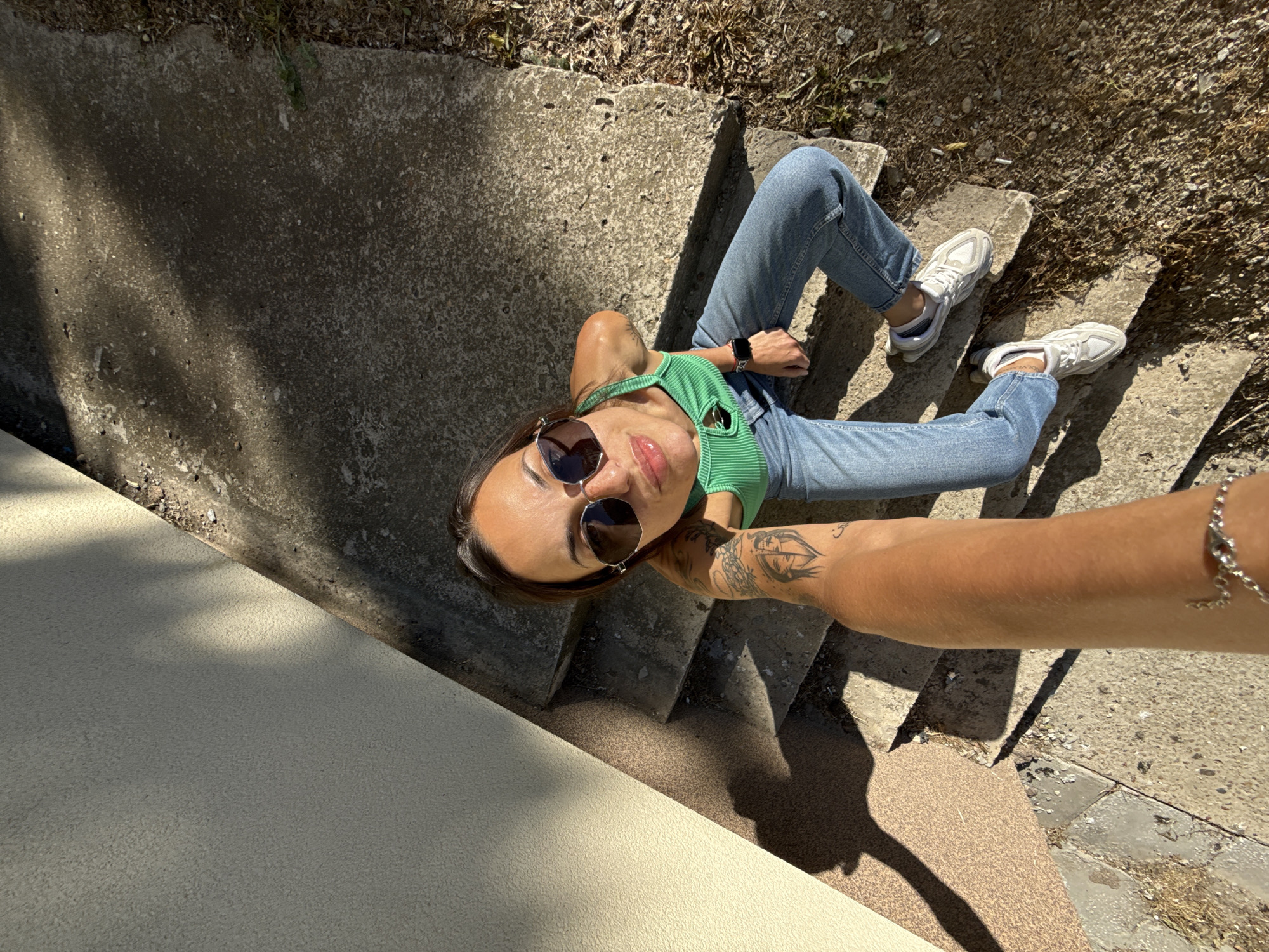 The image depicts a woman taking a selfie outdoors on concrete stairs. She is wearing sunglasses, a green tank top, and light blue jeans. Her feet are clad in white sneakers. She has a watch on her left wrist and a bracelet on her right. A tattoo is visible on her left forearm. The background includes vegetation and a painted wall. The location is likely a public staircase in Pleven, Bulgaria.