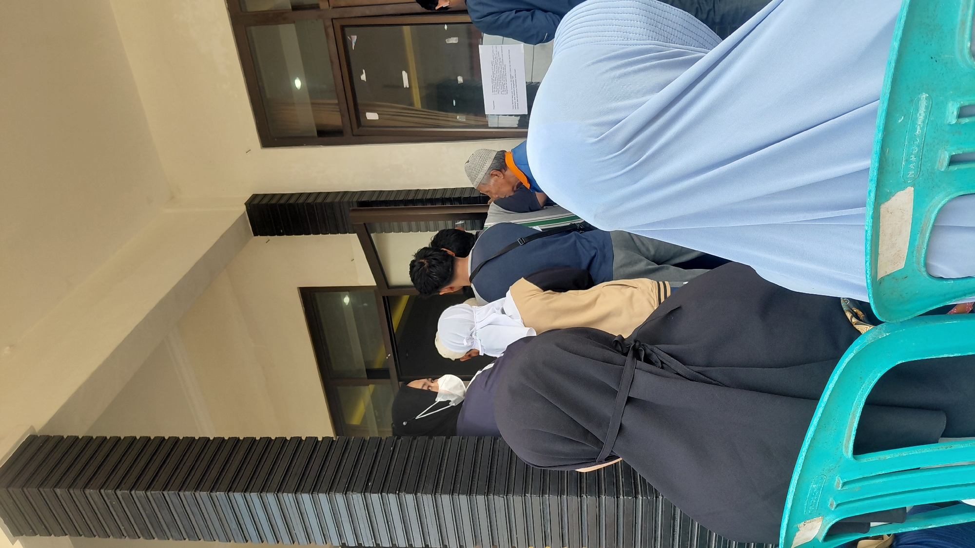 In Pasirmuncang, Indonesia, an outdoor scene depicts a group of people standing in a queue. The individuals are varied in age and attire, with several women wearing hijabs and face masks. One woman in the foreground is dressed in a black hijab. To her right, another woman in a light blue hijab is seated. Further back, a group of men and women are visible, some looking towards a building entrance with glass doors. An older man in a cap and orange jacket is speaking with someone. The environment appears to be a public space, possibly outside a government building or a service center, indicated by the presence of a sign on the door. The architectural style features dark, textured pillars and cream-colored walls. Two bright turquoise plastic chairs are in the foreground, one occupied by the person in the blue hijab.