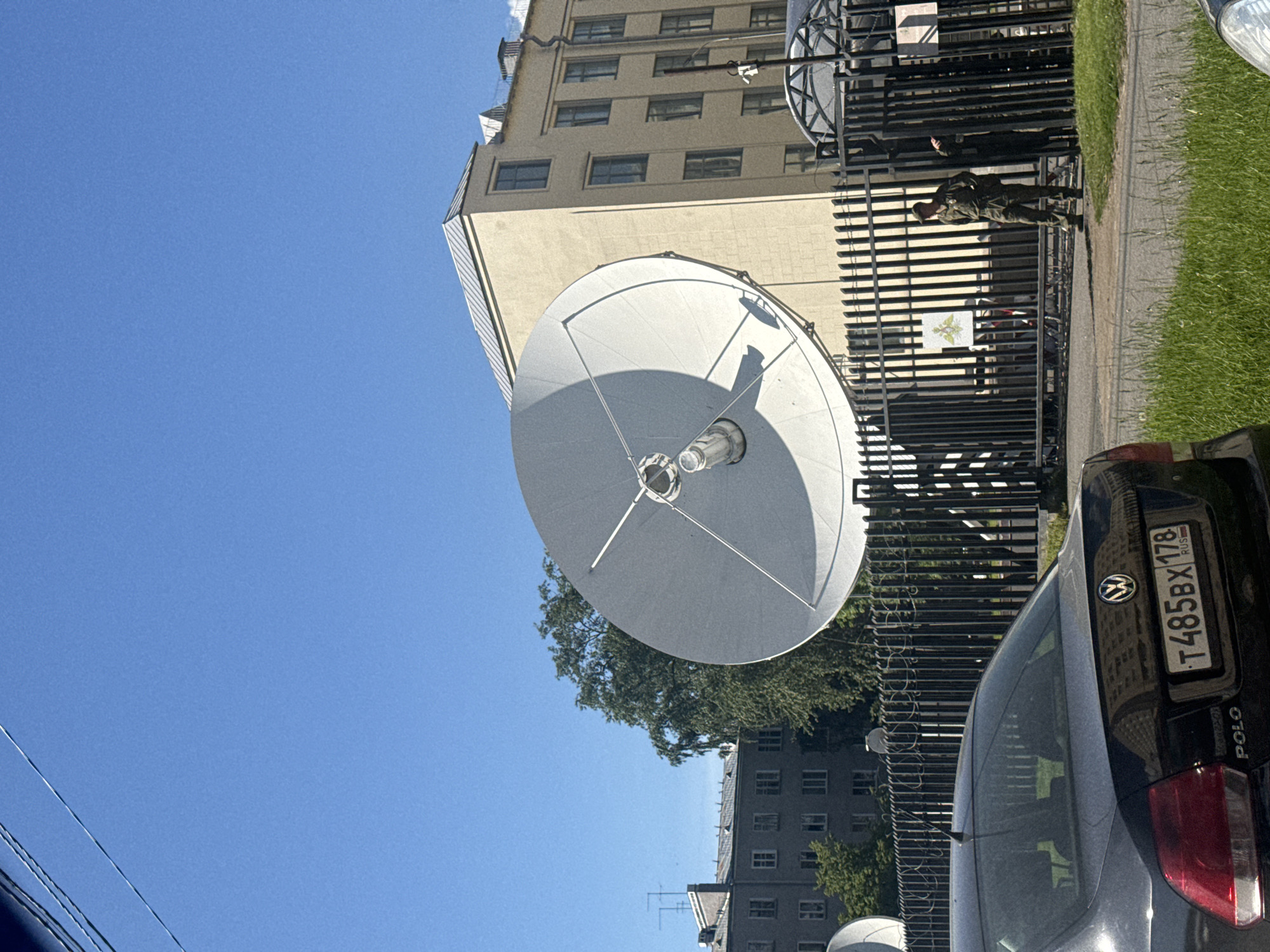 This image captures an urban outdoor scene under a clear blue sky on a bright, sunny day, likely in Saint Petersburg, Russia.
In the left foreground and midground, a large, white satellite dish is prominently featured, casting a distinct shadow. Behind and to the right of the satellite dish stands a multi-story, light-colored building with numerous windows, suggesting an office or institutional building.
A dark metal fence with vertical bars stretches horizontally across the midground, separating a paved area with a strip of green grass from the building complex. A person dressed in a dark olive-green military-style uniform is standing next to this fence, appearing to observe something or someone. Behind the fence, a flag or emblem, which appears to be the coat of arms of Saint Petersburg, is visible.
To the right in the foreground, the rear section of a black Volkswagen Polo car is partially visible. The car bears a Russian license plate, "T485BX 178 RUS," with "178" indicating the Saint Petersburg region. Further in the background on the right, another, darker residential-style building can be seen amidst some trees. The bright sunlight creates sharp shadows, confirming it is daytime. Power lines are subtly visible against the sky in the upper left. The overall scene depicts a static, everyday moment in a city.