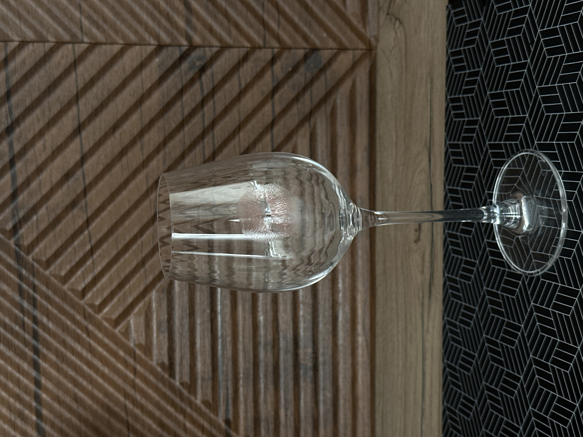 A wine glass rests on two different surfaces. The left portion of the wine glass, including the bowl, is on a wooden surface with a geometric, angled pattern. The right portion of the glass, including the base, is on a black surface covered in a geometric, three-dimensional cube pattern. The background is blurred, but the objects appear to be indoors. The location is Подшивалово, Russia.