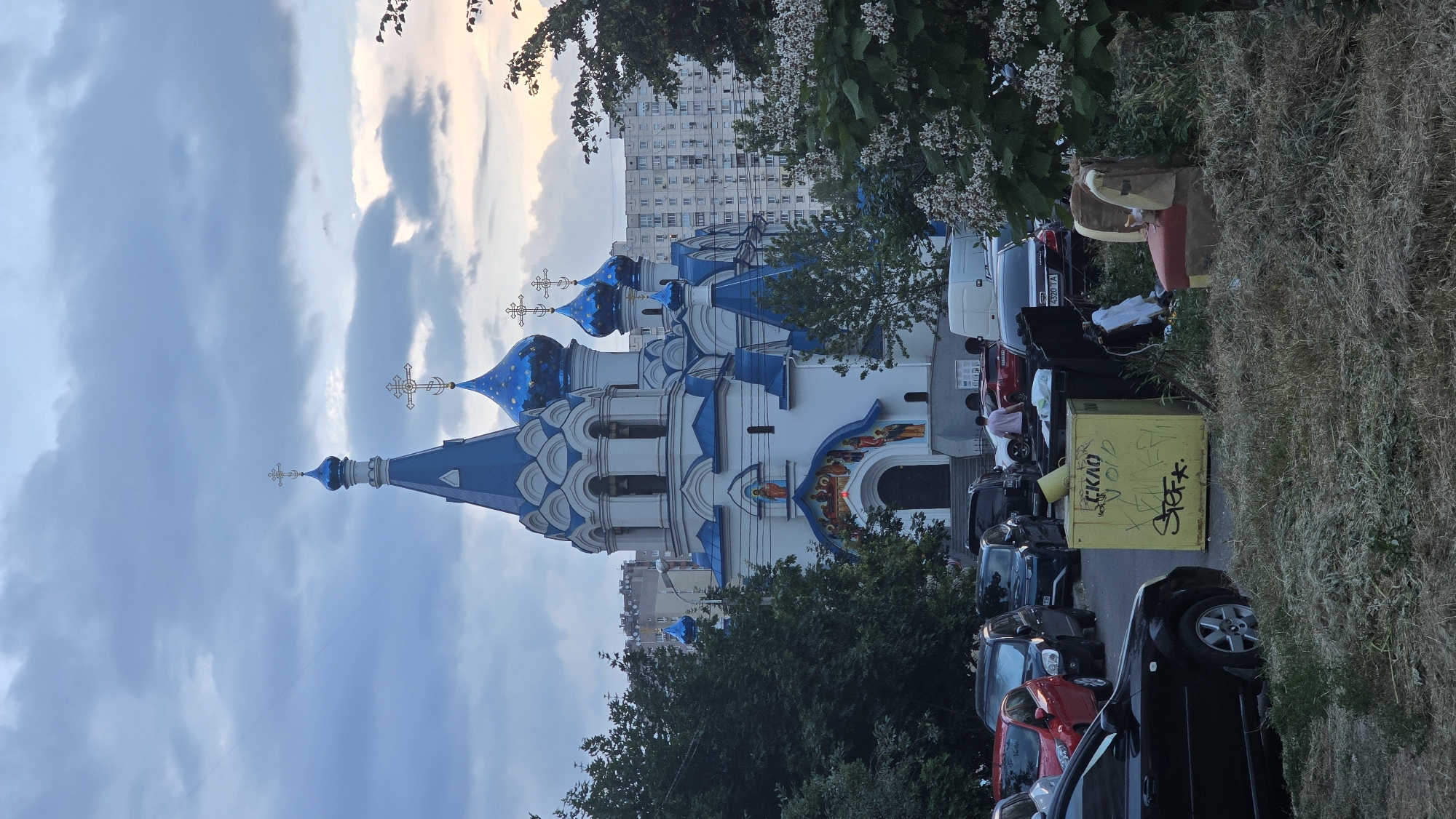 This image captures a view of an Orthodox church with blue domes and white walls, situated in a cityscape. The church is adorned with crosses on its domes. In the foreground, there's a row of parked cars, and to the right, a patch of dry grass and some overgrowth. A yellow dumpster with graffiti is visible among the parked cars.
Behind the church, a multi-story apartment building stands, suggesting an urban environment. Trees and foliage are present, some in bloom with small white flowers, framing the scene. The sky is cloudy with hints of sunset colors, indicating it is likely late afternoon or early evening.
The image appears to be taken from a street level, looking towards the church and the surrounding area. The overall mood is calm, with the urban elements and the church suggesting a place of worship within a residential district. There is graffiti on the yellow dumpster that says "CKLO" and other illegible markings. The license plate on one of the cars reads "4520 TA".