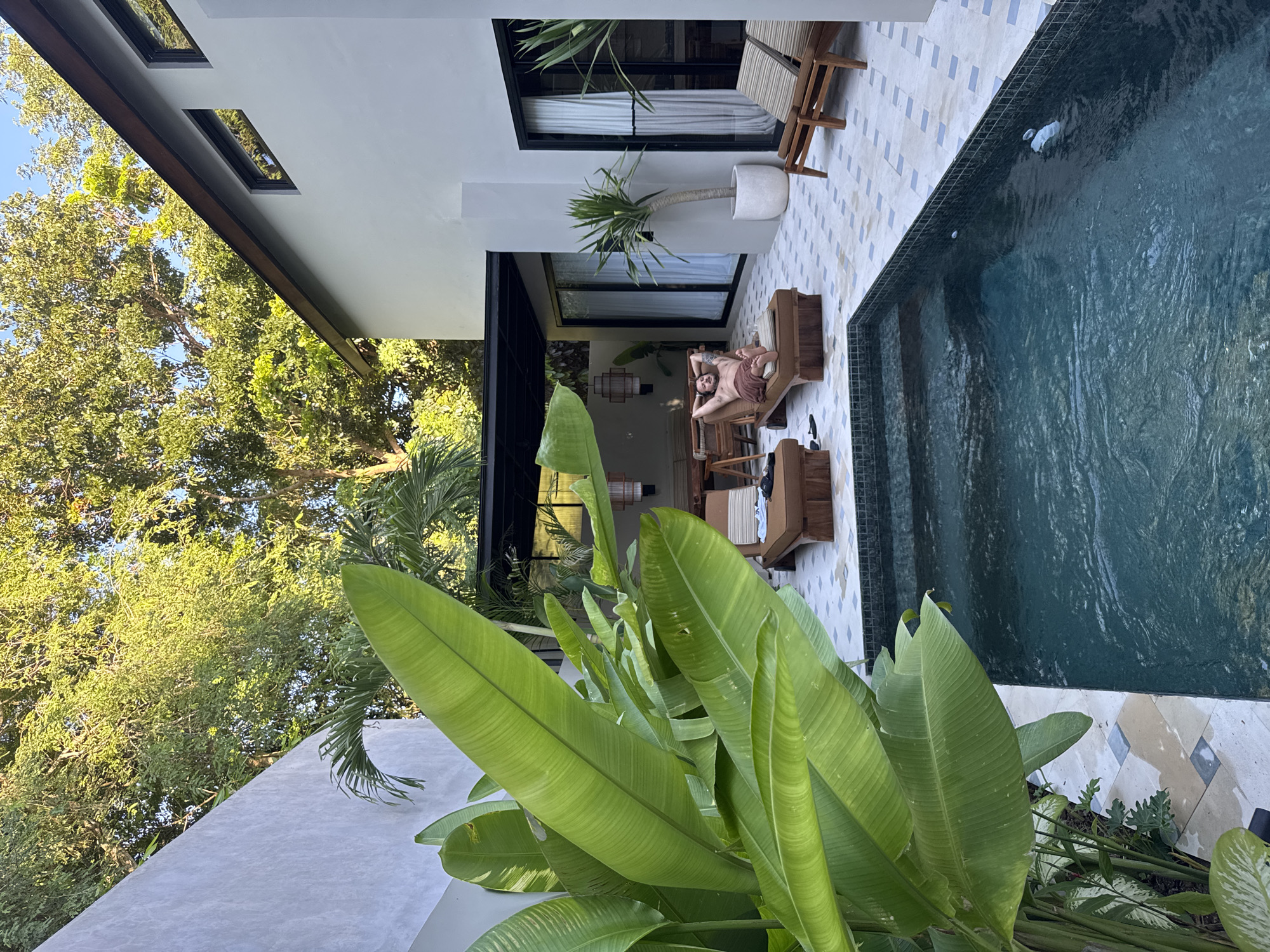 The image depicts an inviting outdoor scene at a modern tropical villa or resort, situated in what appears to be a lush natural environment, consistent with the Kuta Selatan, Indonesia context. The perspective is slightly elevated, looking across a patio area towards a swimming pool and a contemporary building.
In the foreground and left, large, vivid green tropical plant leaves frame the view. A rectangular swimming pool, tiled in dark blue or green, occupies the right side of the frame, showing gentle ripples on its surface. Steps lead into the pool from the patio.
The patio itself is paved with light-colored tiles, featuring a subtle geometric pattern. On the patio, a man is relaxing on a wooden sun lounger. He is lying on his back, wearing a light-colored wrap or sarong around his waist. His hands are resting on his torso, and he appears to be enjoying the sun. Nearby, another wooden lounger and additional outdoor furniture are visible. A potted plant with long, spiky leaves stands near the building.
The building exhibits modern architecture with clean white walls and dark-framed windows or sliding glass doors, which reflect some of the surrounding greenery. A prominent dark-colored roof overhang extends over the patio. Beyond the building and its roofline, dense green trees indicate a rich natural landscape. The bright, clear lighting suggests it is daytime with sunny weather, creating a tranquil and luxurious atmosphere. No text is visible in the image.