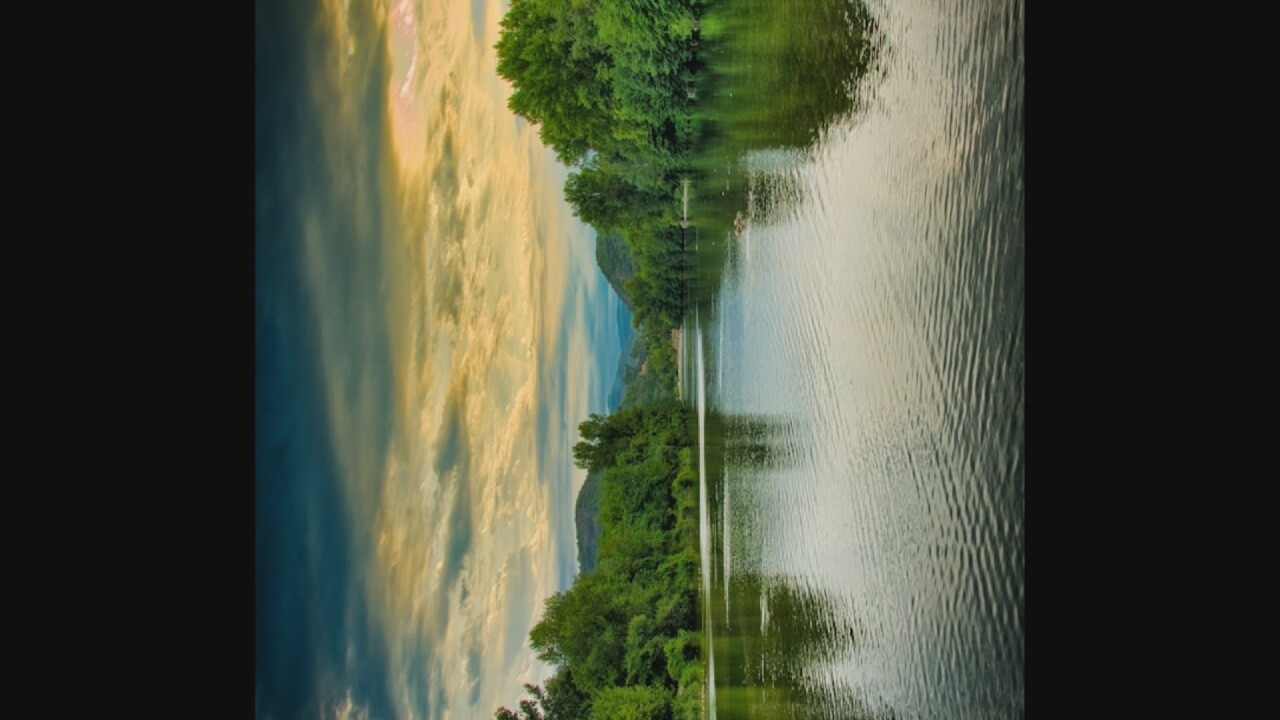 The image depicts a wide, calm river flowing through a lush, green landscape. The riverbanks are densely lined with trees, with hills visible in the background. The sky above is filled with dramatic, cloudy formations, with hints of yellow and orange light suggesting either sunrise or sunset. The water reflects the trees and the sky, creating a serene and natural scene. There are no people or man-made structures visible in the image, indicating a remote or natural setting. The location is not Moscow, Russia.