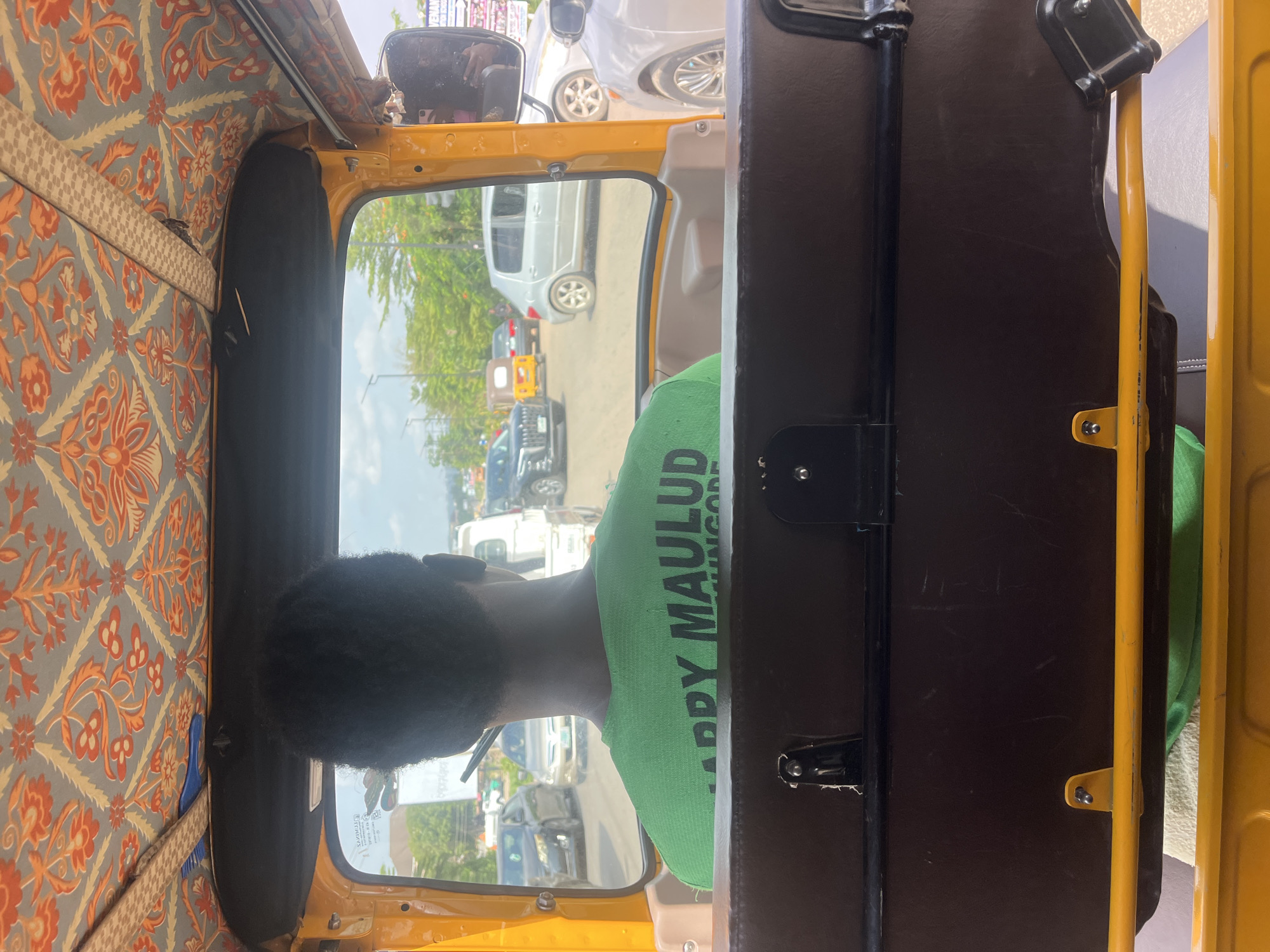 The image shows the interior of a yellow "keke" or auto-rickshaw, a common mode of transportation in many cities, possibly in Abuja, Nigeria. A Black person with short, curly hair and wearing a green t-shirt with black text that reads "HAPPY MAULUD" sits facing forward. The interior of the keke has orange and grey patterned fabric on the roof and side. Outside the window, there are vehicles on a road and some trees.