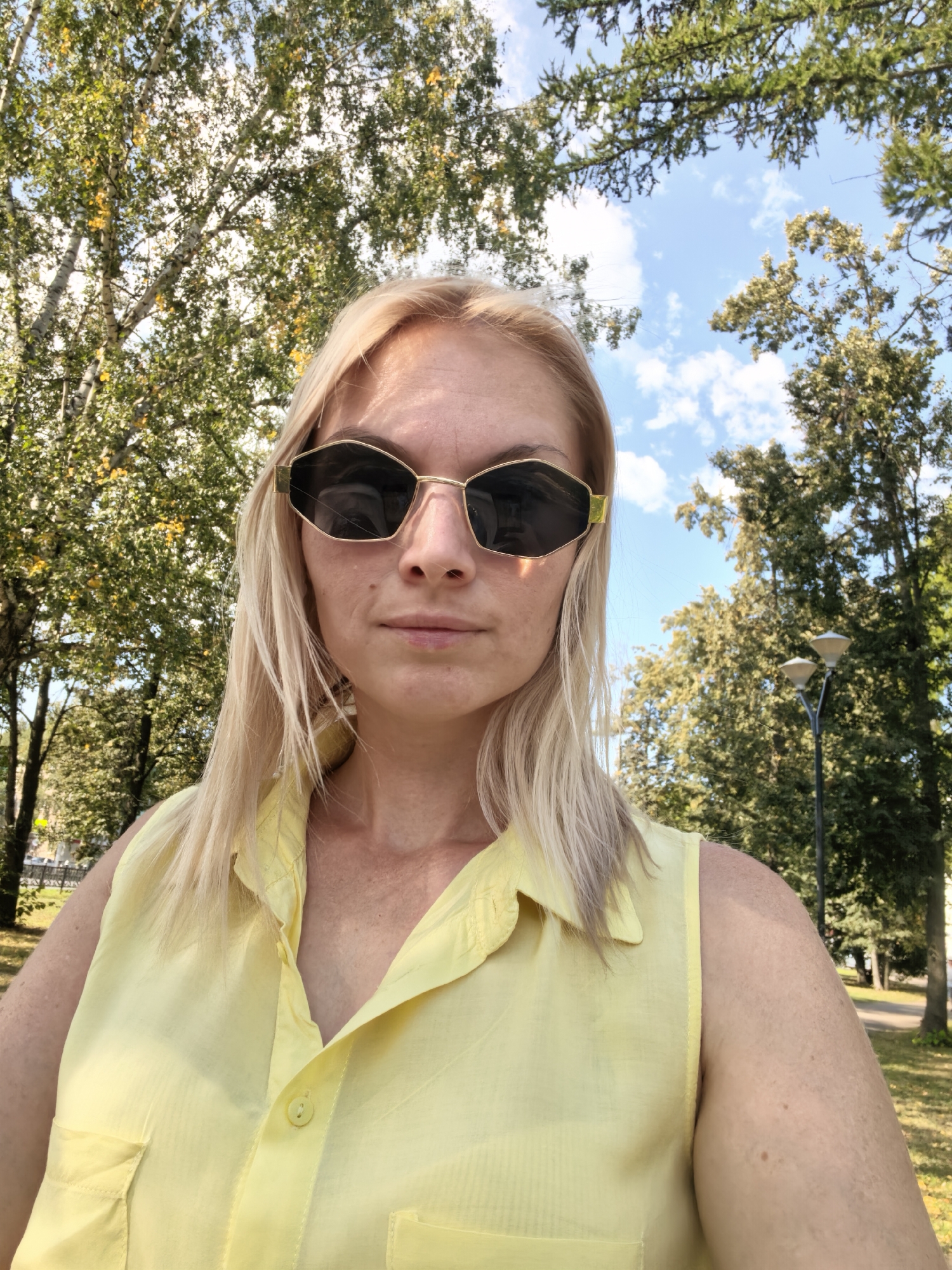 A woman with blonde hair and sunglasses is taking a selfie outdoors. She is wearing a yellow sleeveless shirt. The background consists of trees and a blue sky with clouds. The image appears to have been taken in a park or wooded area. There are no specific landmarks visible that would indicate the city of Sterlitamak, Russia.