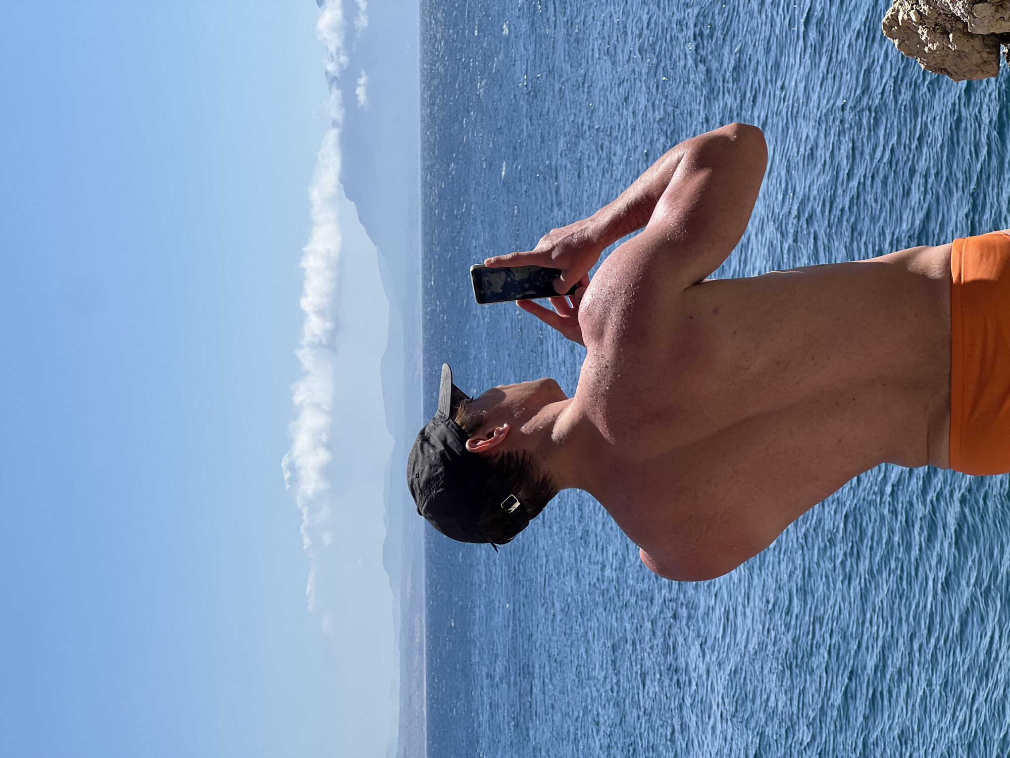 A shirtless person wearing an orange bathing suit and a dark baseball cap stands on a rocky outcrop overlooking the sea. They are holding a smartphone in their hands and appear to be taking a picture or recording a video of the distant, hazy coastline under a bright blue sky with scattered clouds. The sea is a deep blue with visible waves. The overall scene suggests a sunny day by the coast in Terrasini, Italy.