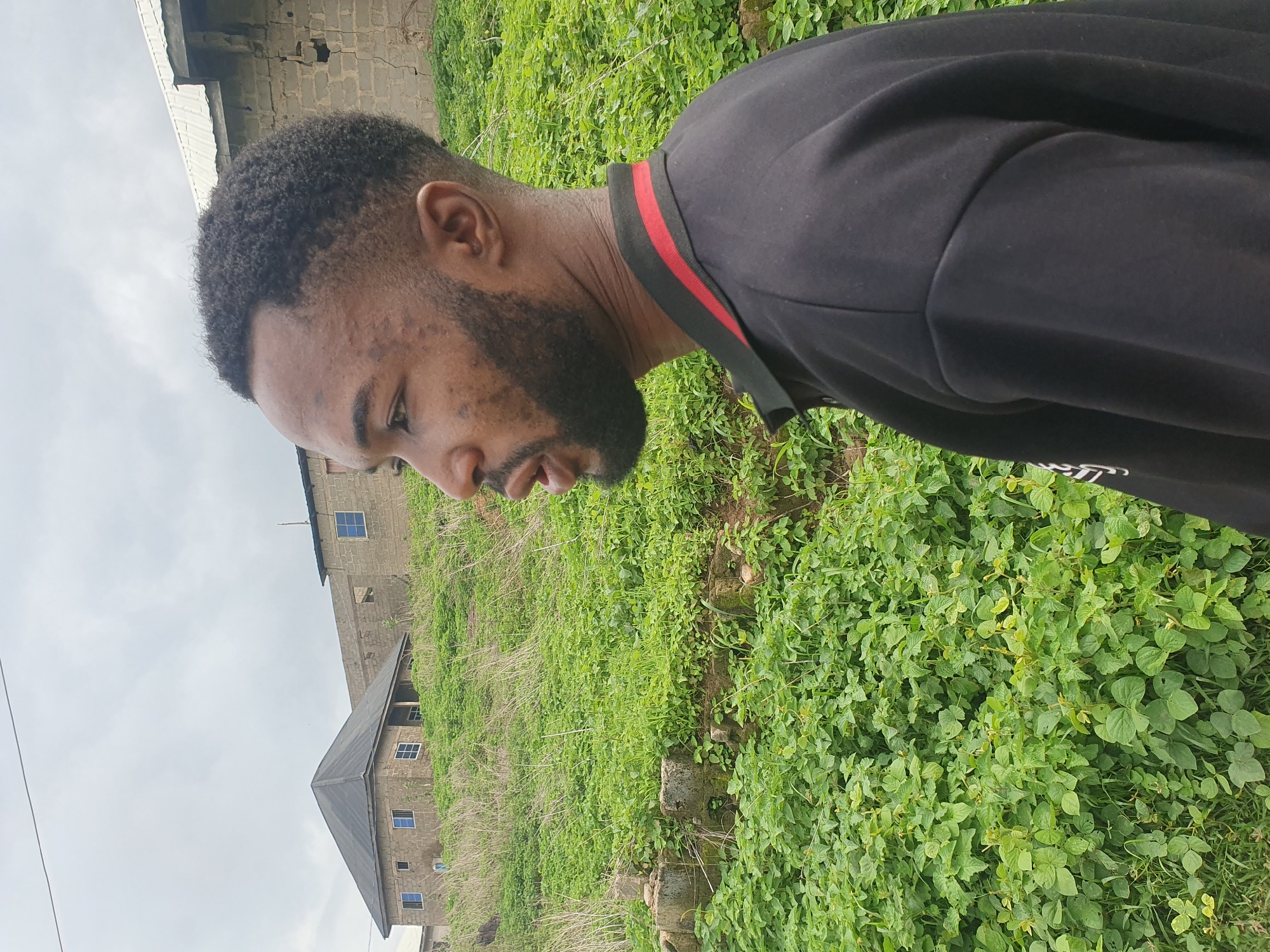 A young man with dark skin and short, curly hair, wearing a black polo shirt with red and black striped trim, is captured in a profile view facing left. His mouth is open, as if speaking or reacting. He has a beard and light acne scarring on his face. He stands in an outdoor setting characterized by lush green vegetation and some older, unfinished brick buildings in the background. The landscape appears somewhat rugged and overgrown, with dry grasses interspersed with the greener foliage. In the distance, one building is a two-story structure with a dark, sloped roof and visible windows, while another is a more rudimentary construction with exposed brickwork. The sky is overcast, suggesting a cloudy or potentially rainy day. The scene is likely situated in an area undergoing development or where construction is ongoing, indicative of a possibly rural or developing urban periphery within Lokoja, Nigeria.