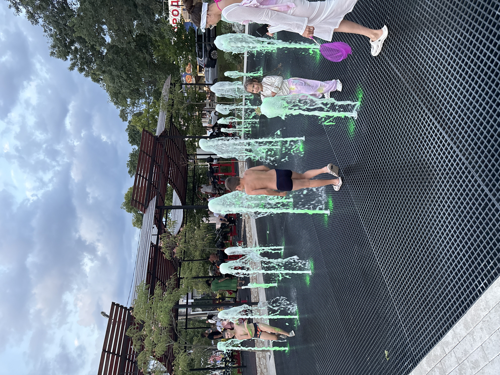 The image depicts a lively scene at a wet playground with multiple water fountains erupting from a dark, grated surface. Several children are actively engaged with the water, with one boy in swim trunks running through the spray and another, also in swim trunks, standing near a large fountain. A young girl in colorful leggings is smiling as she interacts with a fountain, and a woman in a white dress and pink top, wearing a headscarf, walks past holding a purple net. In the background, there are covered seating areas, trees, and buildings, suggesting an outdoor public space in a city. The sky is overcast with dramatic clouds. The water jets are illuminated with a green light, creating a vibrant effect. Based on the signage in the background, which includes Cyrillic script and the word "РОД", the location is likely Novorossiysk, Russia.