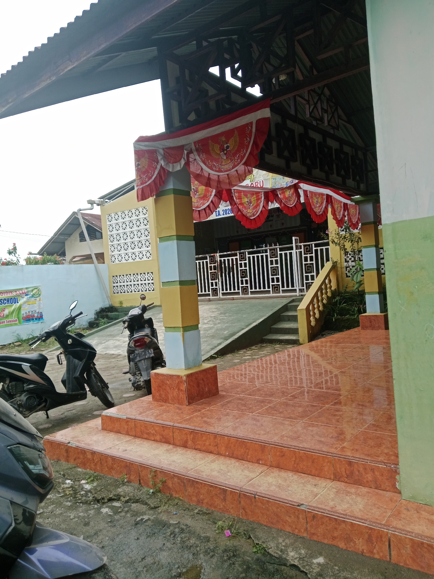 The image depicts the exterior of a building, likely a school, in Galang Kota, Indonesia. Two motorcycles are parked on a concrete surface in front of a set of steps leading up to a covered entrance. The entrance is decorated with red and white pennants featuring the Garuda Pancasila symbol. A light yellow building with white patterned window blocks is visible behind the entrance. The overall environment appears to be slightly overcast.