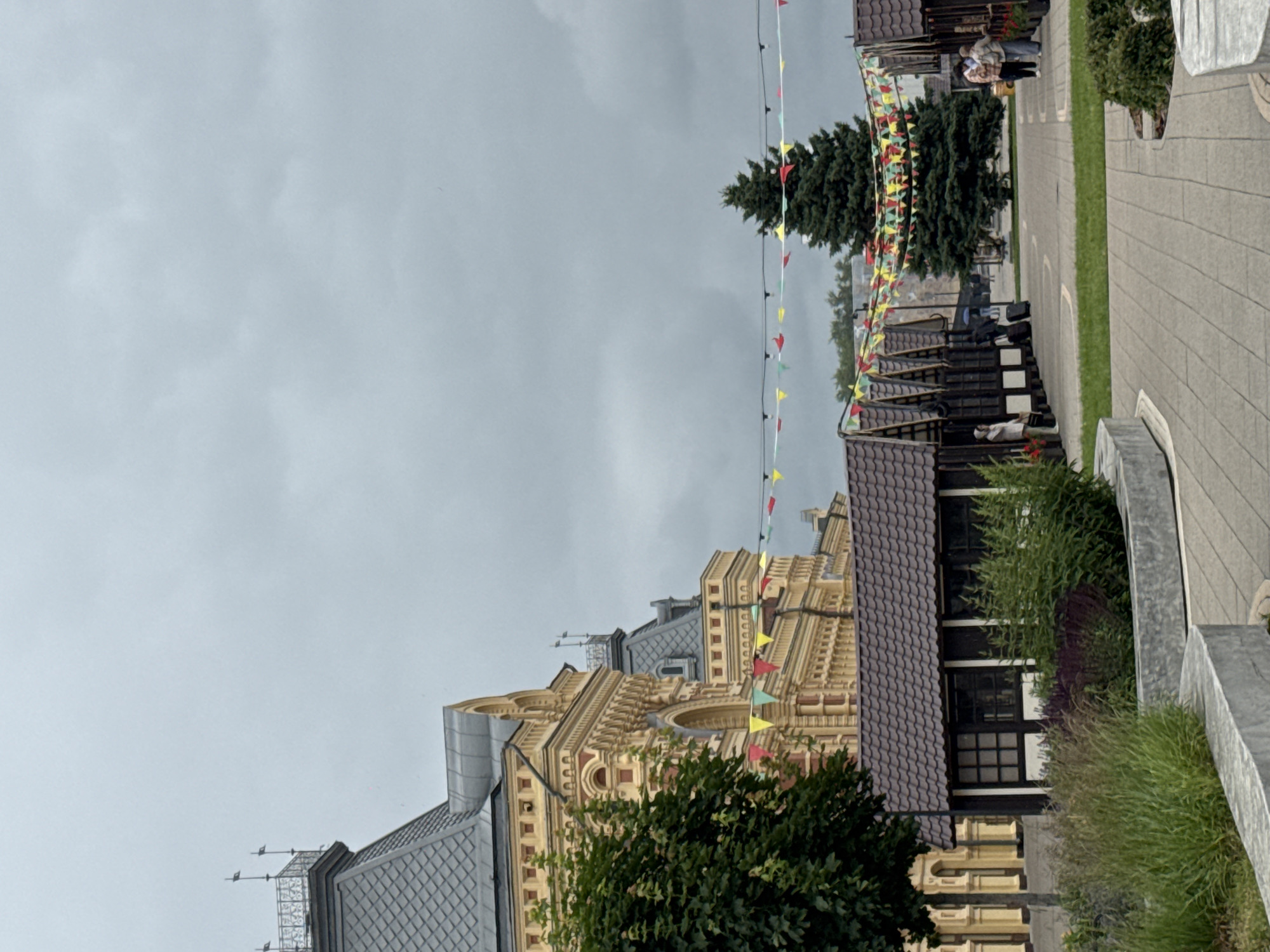 This image shows a view of the Nizhny Novgorod Kremlin on a cloudy day. In the foreground, there are several small, dark wooden stalls with brown tiled roofs, set up in a row. A string of colorful pennant flags is strung between the stalls and across the pathway. Behind the stalls, a large, ornate yellow building with decorative white accents and a dark grey roof is visible. A large evergreen tree stands in the middle ground, with more stalls visible to its right. A few people are seen walking in the background, some with luggage. The ground is paved with light-colored square stones. The overall scene suggests a marketplace or festival within the Kremlin grounds.