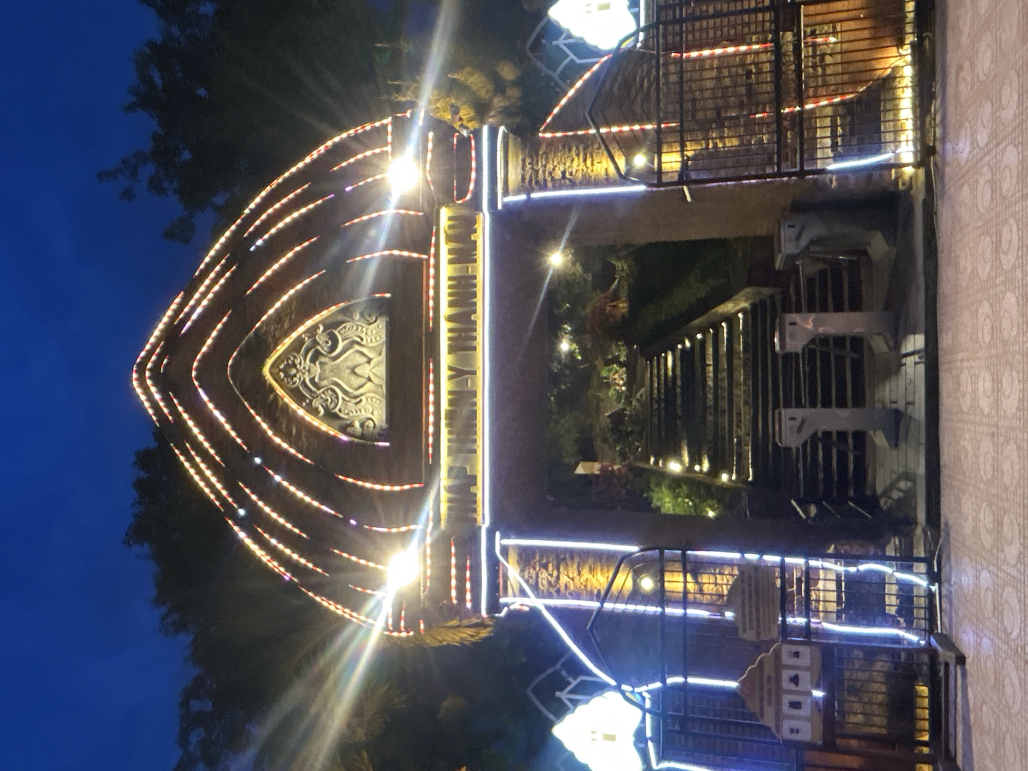 The image shows the entrance to a site, potentially a tourist attraction, in the city of Bac Nha Trang Ward, Vietnam. The entrance is marked by a large, ornately decorated archway, illuminated with numerous lights. There is signage on the archway written in Vietnamese. Beyond the archway, a set of steps leads downwards. The area is surrounded by trees and foliage. In the foreground, there is a railing with decorations and turnstiles. The sky is a deep blue, suggesting it is either dusk or dawn. The ground appears to be paved with a patterned tile.